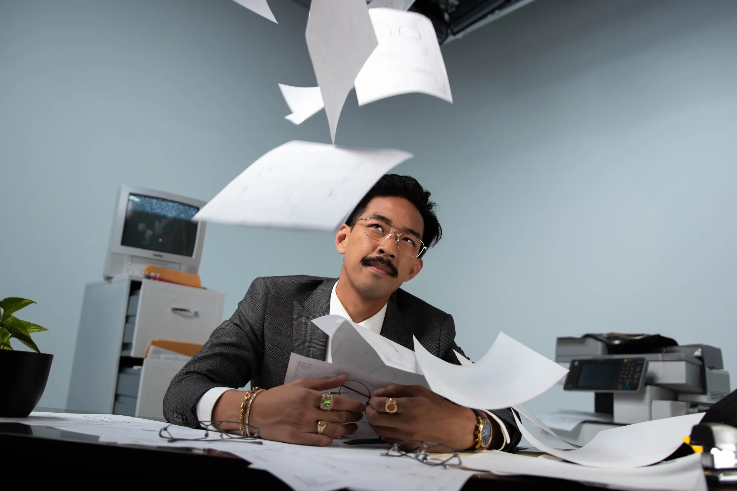 A man with glasses and gold jewelry, including rings, a watch, and bracelets, is sitting at a cluttered desk with papers flying around him. He appears to be frustrated or overwhelmed while working in an office with a computer, printer, monitor, and filing cabinet.