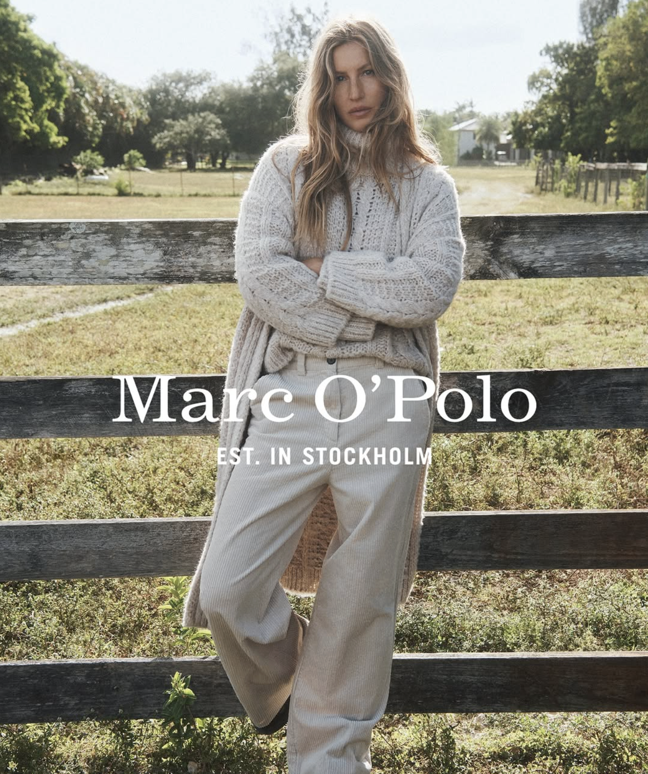 A woman with long wavy hair wearing a cream-colored knitted sweater and light-colored pants, standing outdoors by a weathered wooden fence with a field and trees in the background.