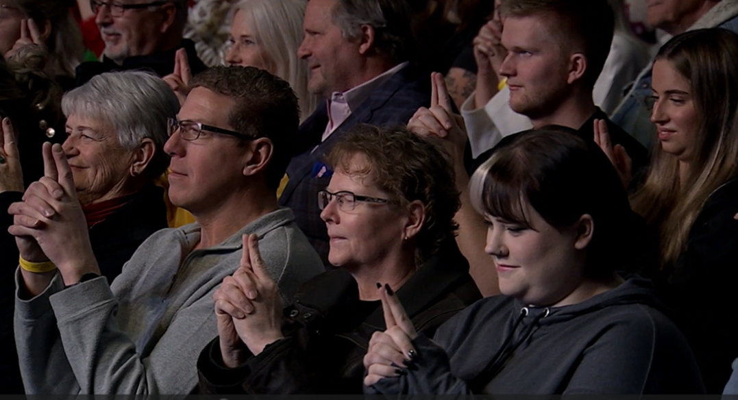 Audience members sitting in an auditorium, some holding up their index fingers as if to indicate 'number one'.