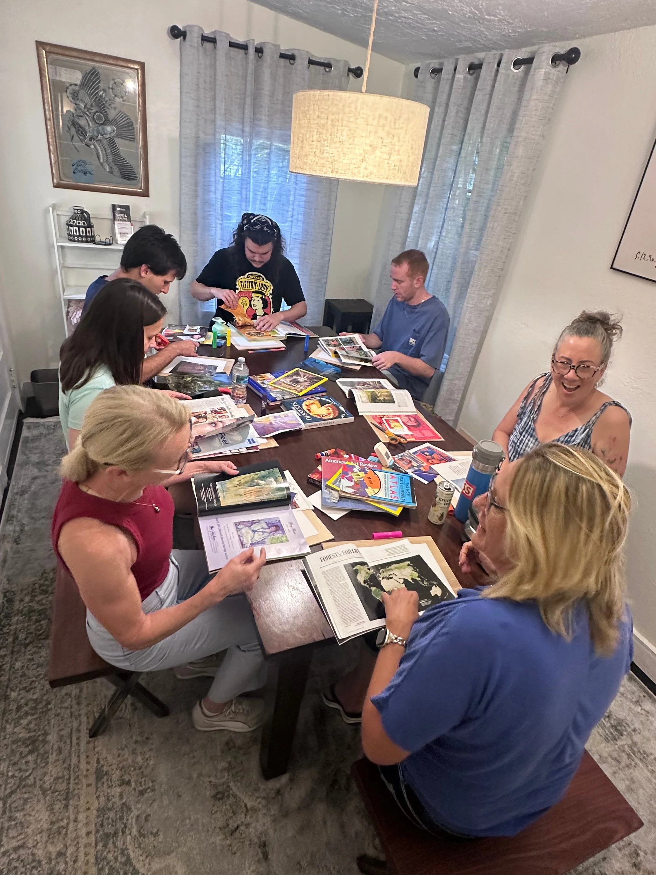 People gathered around a table looking at magazines and books, with some engaged in conversation and others reading.