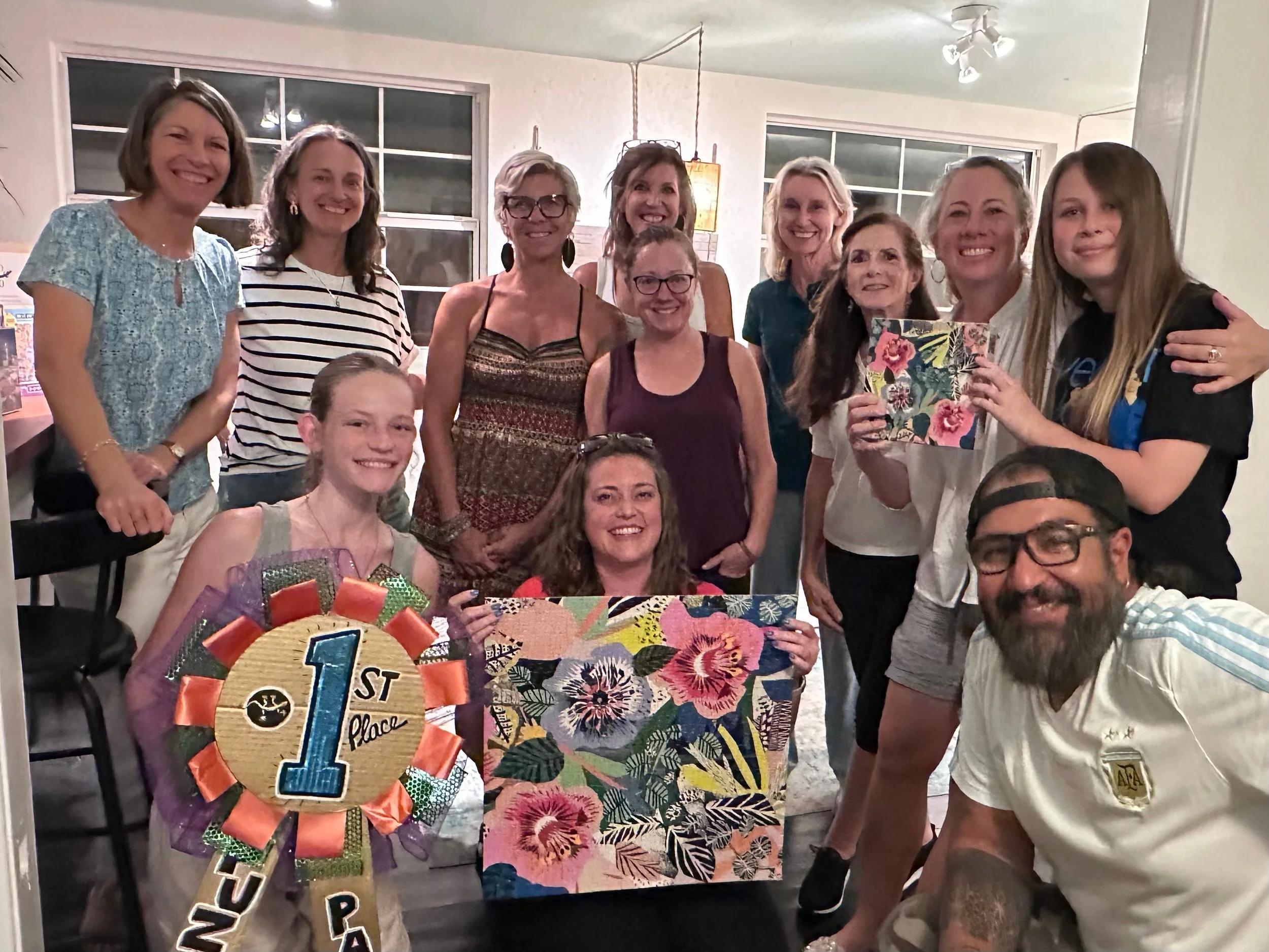 Group of people celebrating a first-place achievement at a party, holding a floral-themed sign and wearing casual attire, with several smiling and posing for the photo.