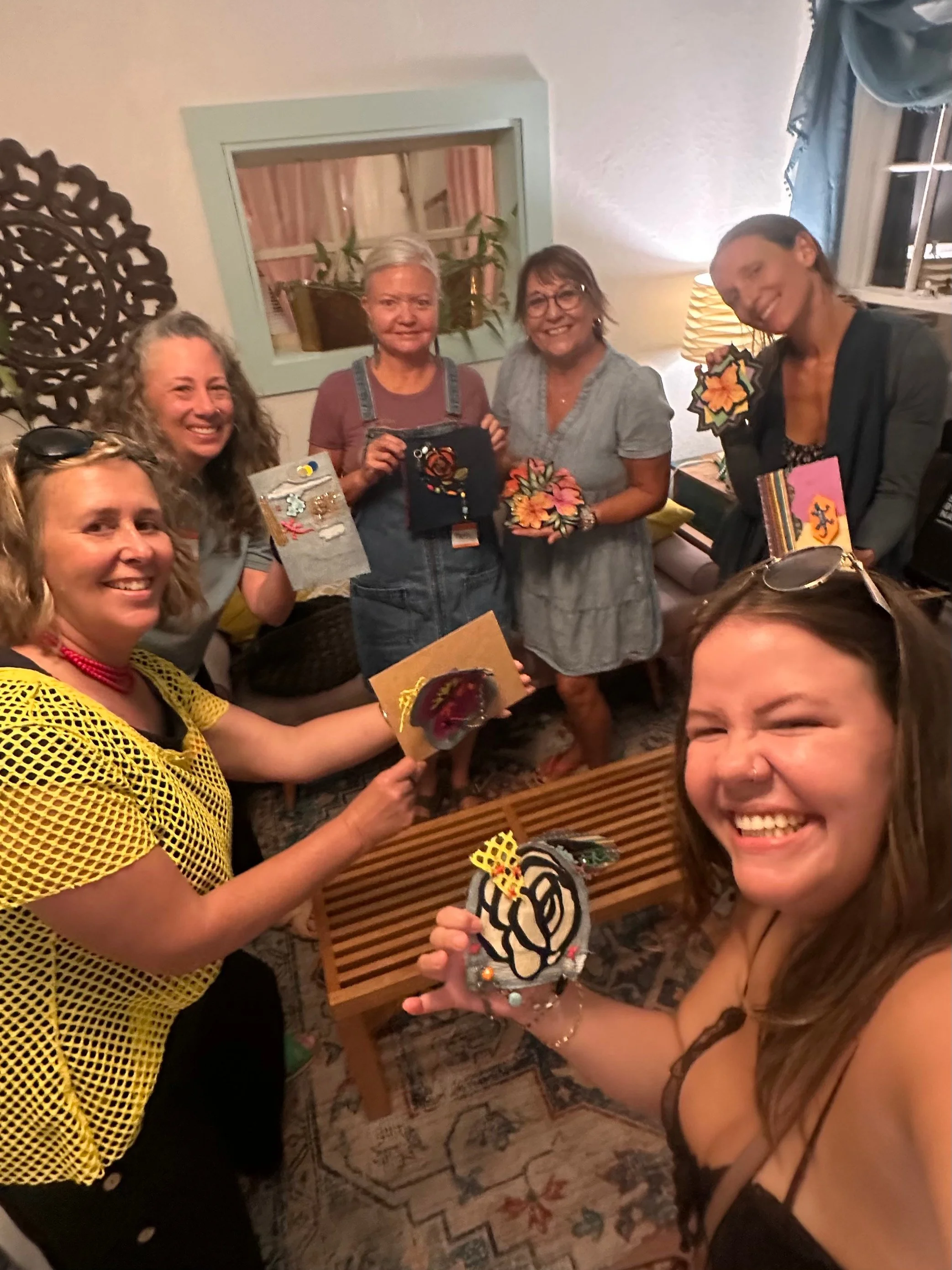 A group of women smiling and holding craft projects in a warmly lit indoor setting.