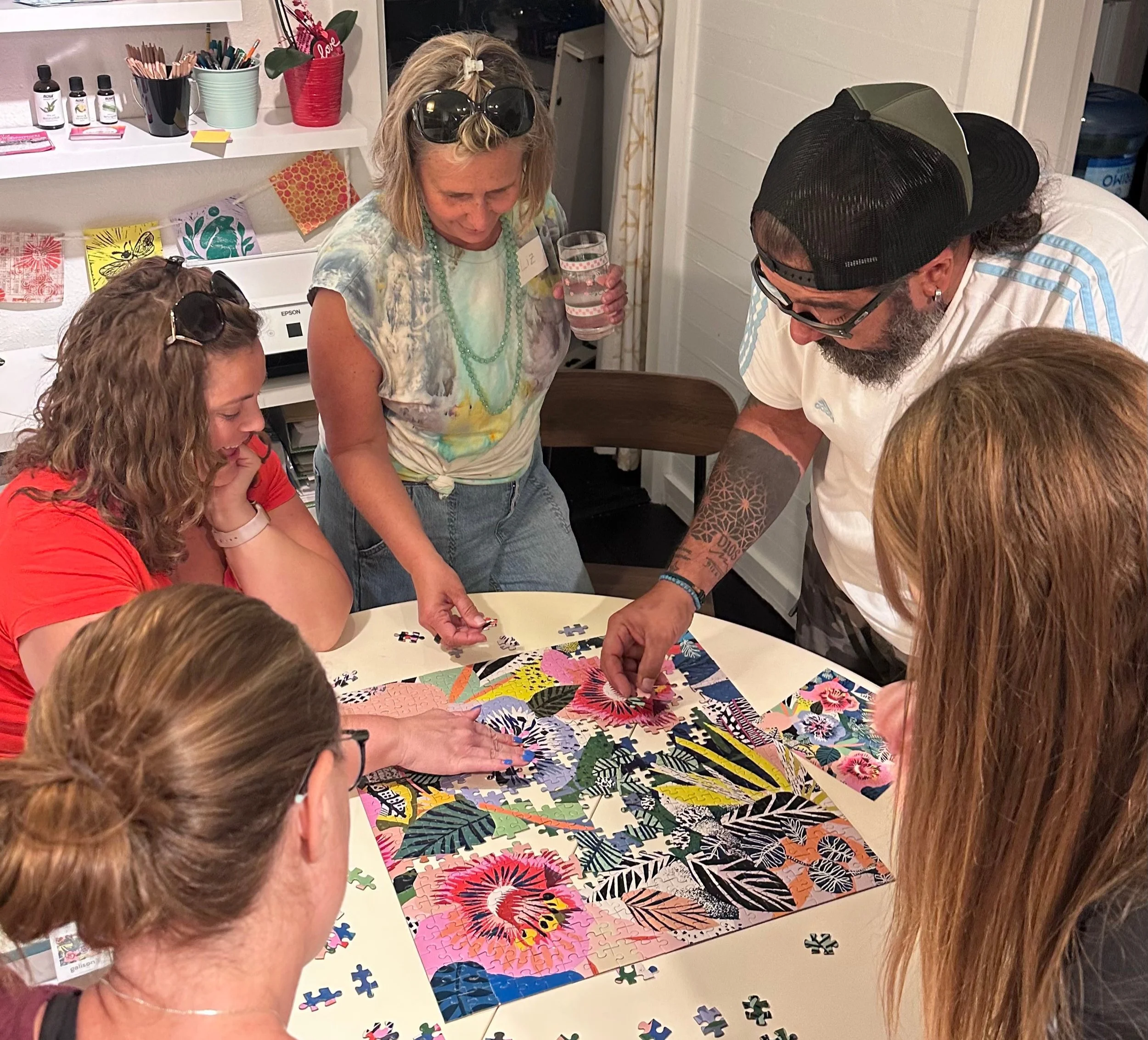 Group of people working on assembling a colorful jigsaw puzzle on a table in a room decorated with artwork and plants.