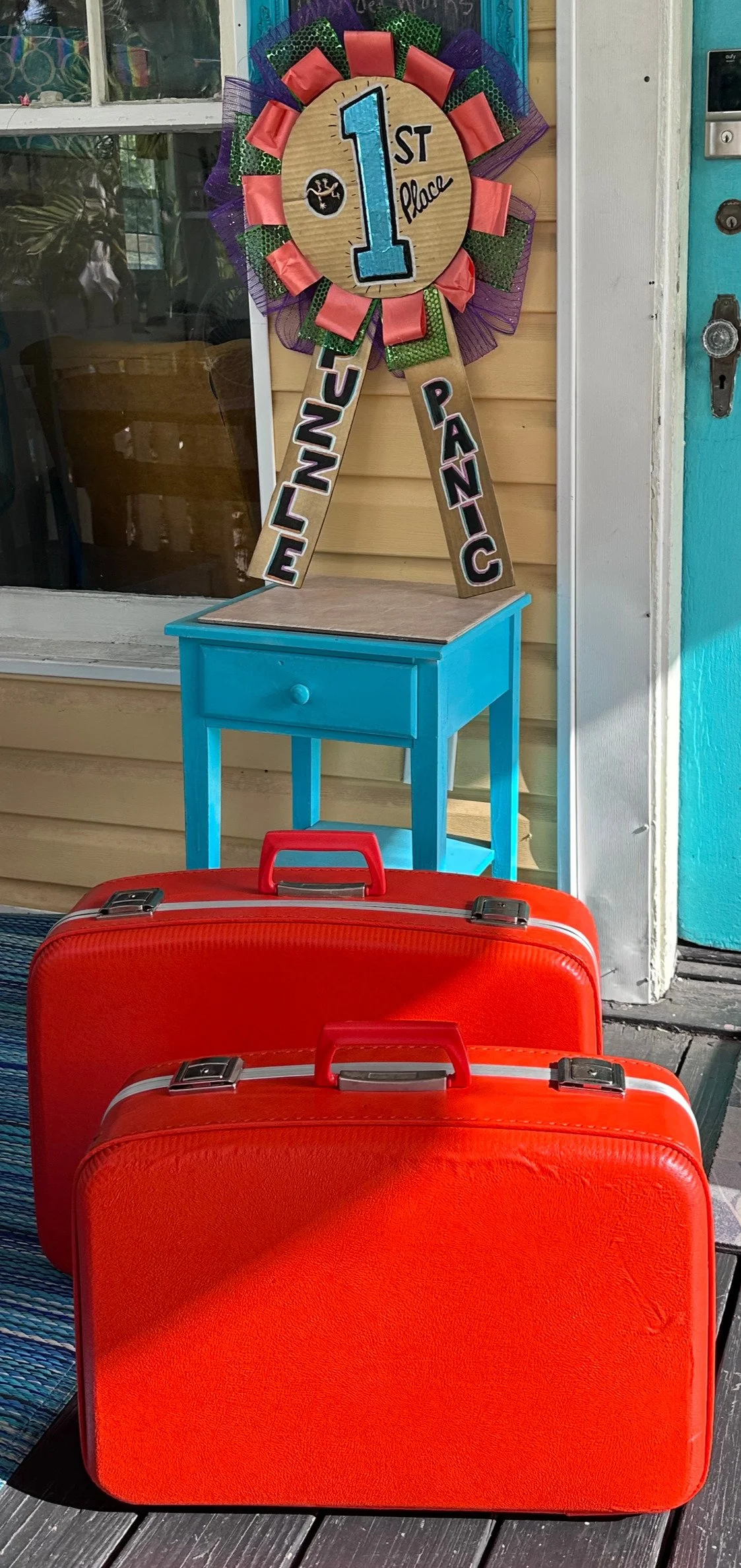 Decorative first place ribbon with colorful rosettes, black lettering spelling "1st Place," and the words "Lineup Panics" on two hanging wooden strips, placed on a small blue table outside near red suitcases.