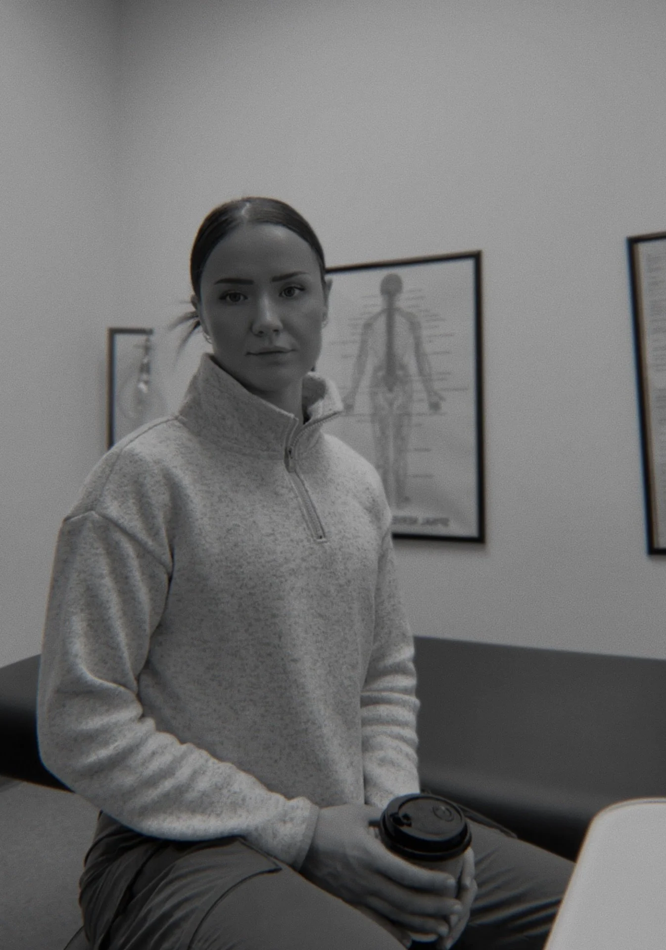 A woman with dark hair tied back, wearing a light gray quarter-zip pullover, sitting in a room with framed anatomical diagrams on the wall, holding a coffee cup on her lap, with a laptop in front of her.