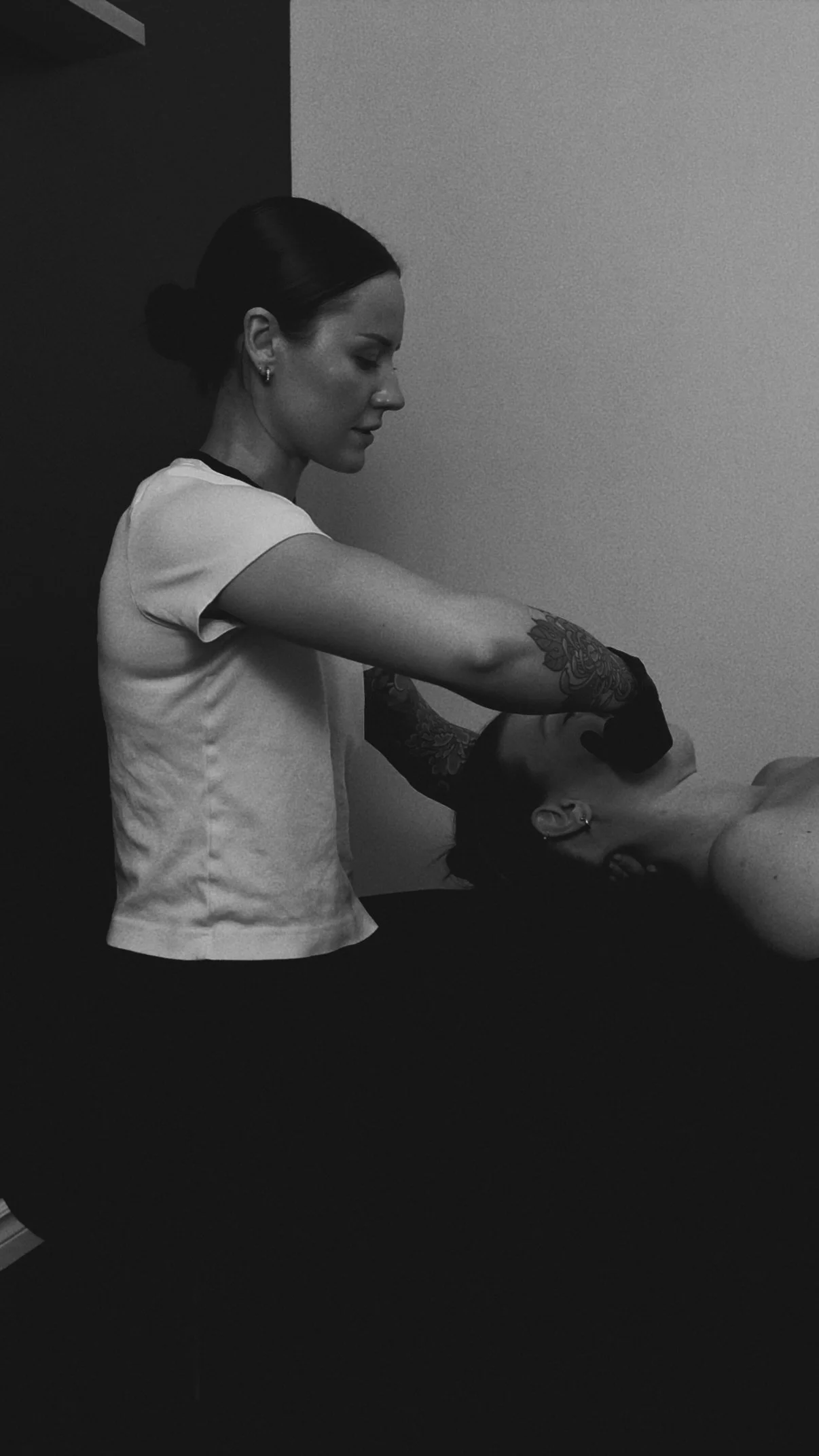 A woman with dark hair in a bun, wearing a light-colored shirt and earrings, is leaning over a person who is lying down with their eyes closed. The woman appears to be performing a facial or skincare treatment.