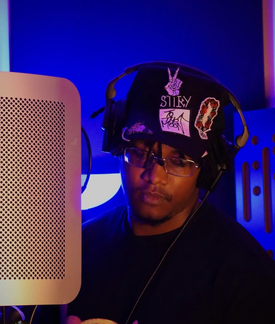 A man with glasses wearing a black beanie with patches, headphones, and a black shirt, recording in a music studio with a colorful, blue-lit background.