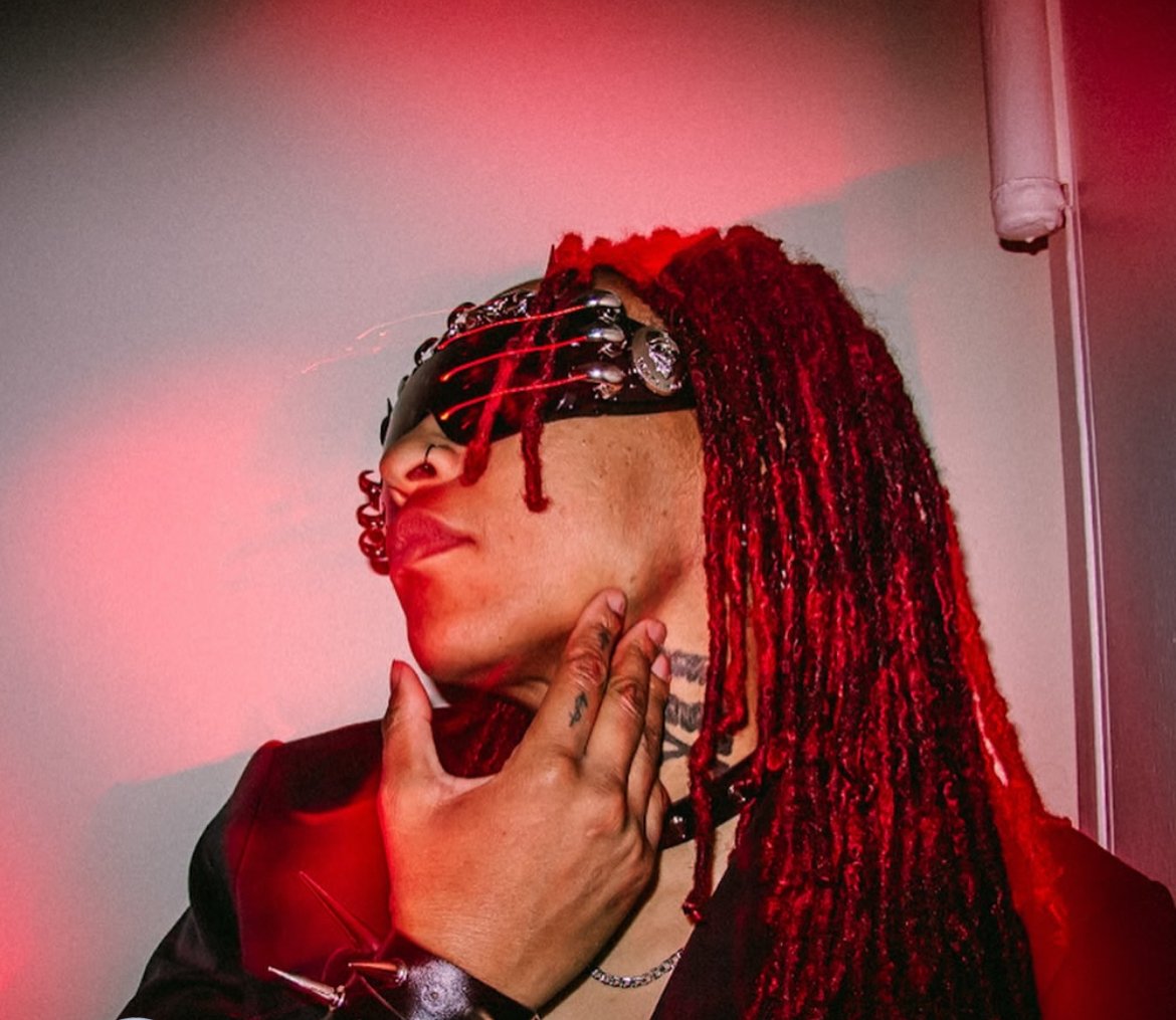 Person with red dreadlocks wearing dark sunglasses, touching their neck, in a room with pink lighting.