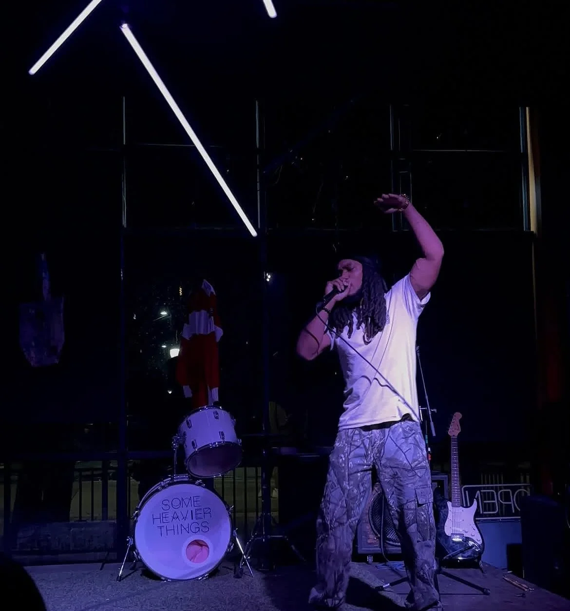 Musician with dreadlocks singing into a microphone on stage with a drum set and electric guitar in the background, in a dark environment with neon lighting.