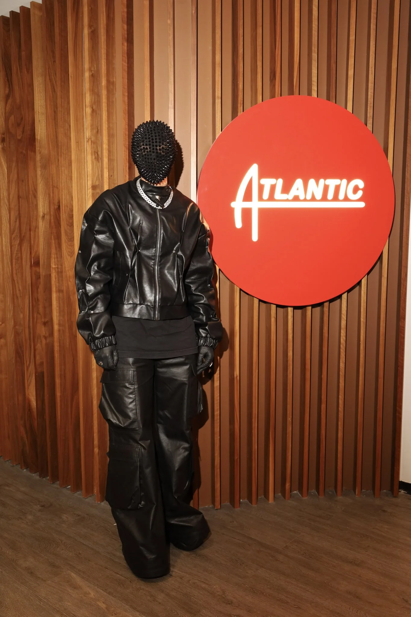 A person dressed in all black, wearing a spiked mask, leather jacket, gloves, and pants, standing in front of a wooden wall with a large red circular sign that reads 'ATLANTIC' in white neon lights.