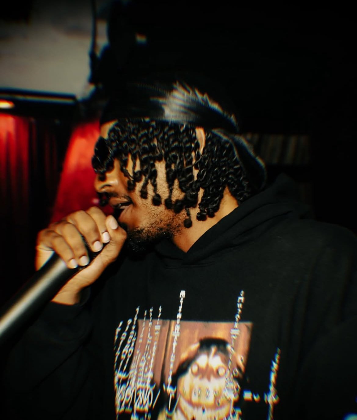 A man with curly hair and wearing a black cap, holding a microphone close to his mouth, possibly performing or singing, wearing a black hoodie with a graphic print.