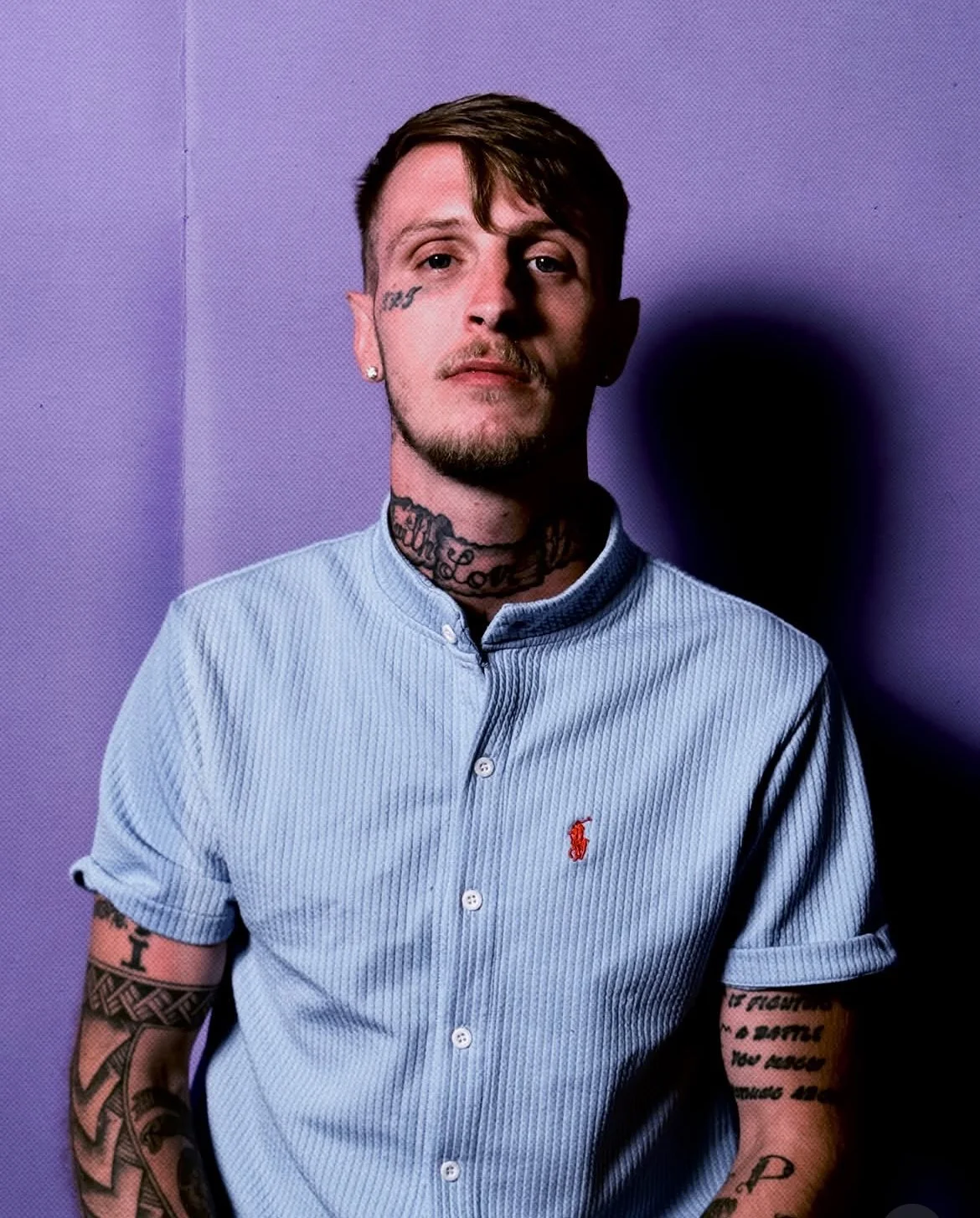 A young man with tattoos on his neck and arms stands against a purple wall. He has short brown hair, a goatee, and is wearing a light blue, short-sleeved, button-up shirt with a red logo.