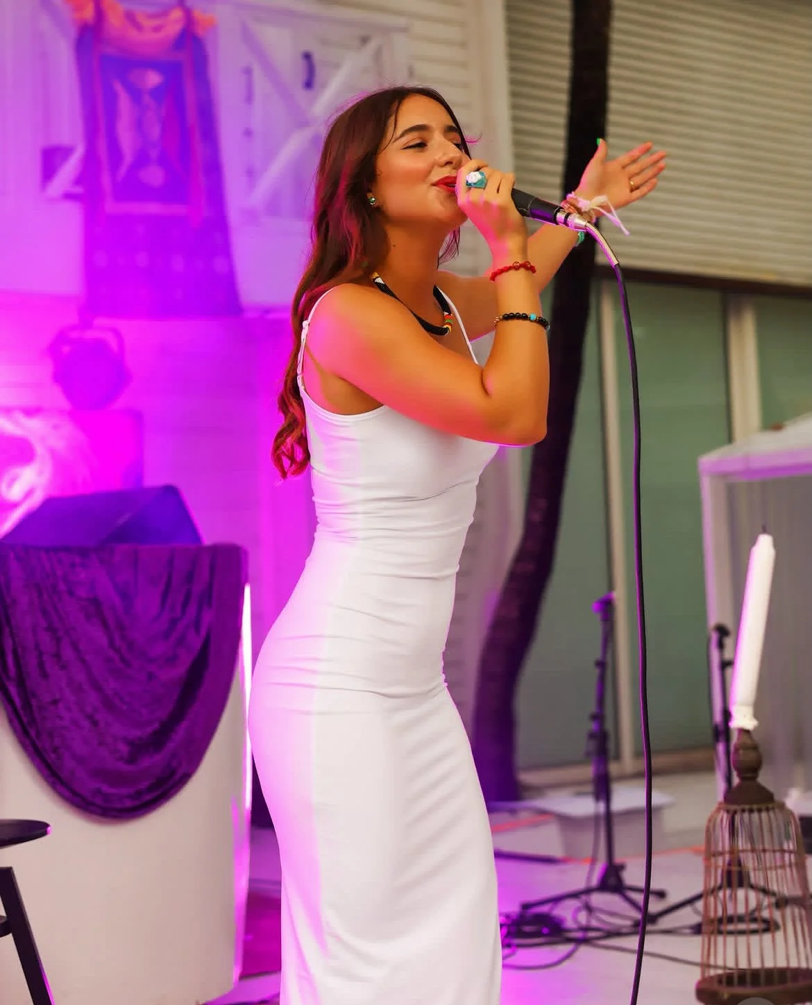 A young woman singing into a microphone with her eyes closed, wearing a white sleeveless dress, with colorful accessories, in a decorated indoor setting with purple lighting.