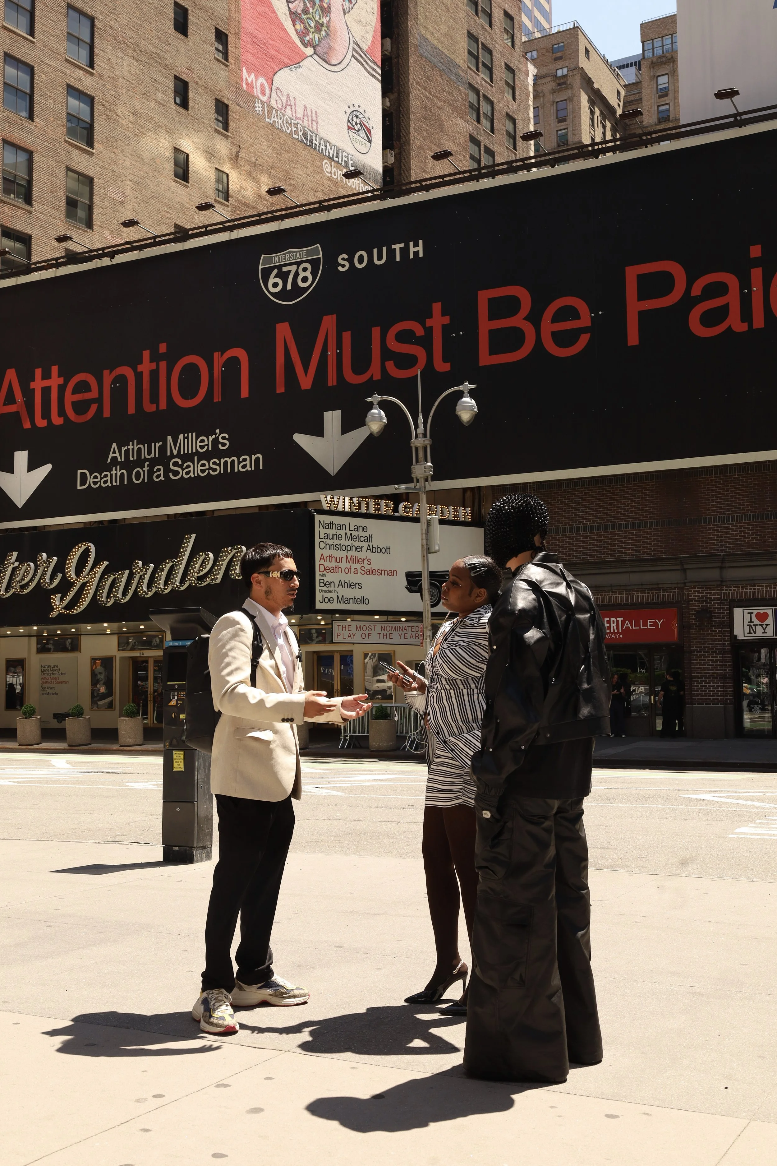 Three people standing on a city sidewalk in front of a large black billboard advertisement. The billboard features a large, partially visible face illustration and text with directions to Interstate 678 South and a note about Arthur Miller's play. One person, dressed in a light-colored suit, is facing two women, one in a striped top and pants, and the other in a leather jacket, who are facing each other and engaging in conversation. There are buildings, a street lamp, and more signs in the background.