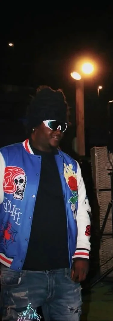 A man with sunglasses and dreadlocks wearing a colorful varsity jacket with skull and floral designs, standing outdoors at night near a lit street lamp.