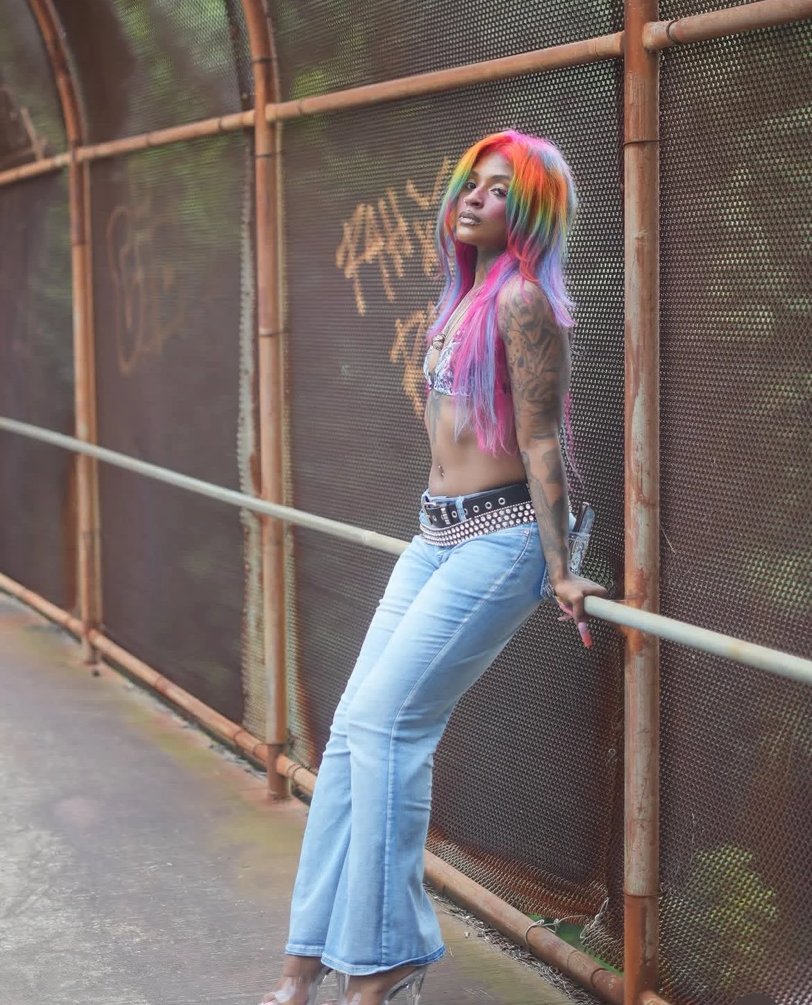 A woman with rainbow-colored hair wearing a patterned crop top, light blue flared jeans, and high heels, leaning against a railing next to a rusted metal fence with graffiti in the background.