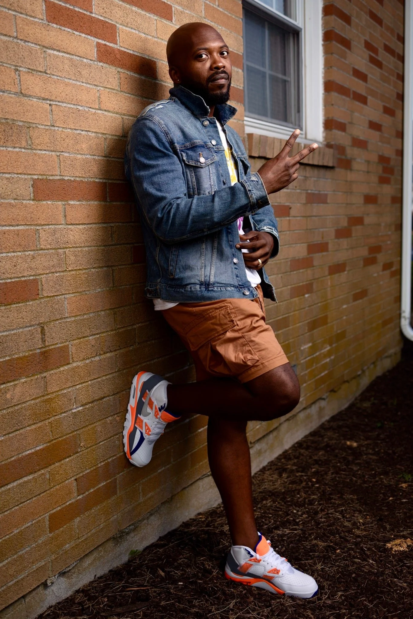 A man leaning against a brick wall outdoors, wearing a denim jacket, orange shorts, and white athletic shoes with orange and navy accents, making a peace sign with his right hand.