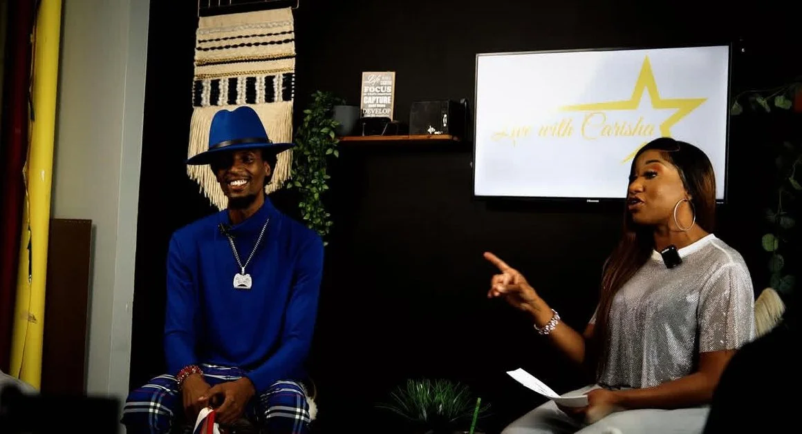 Two people in a conversation, one man smiling wearing a blue hat, shirt, and plaid pants, and a woman speaking while pointing, sitting in front of a monitor with a star and text, in an indoor setting with decorative wall hangings.