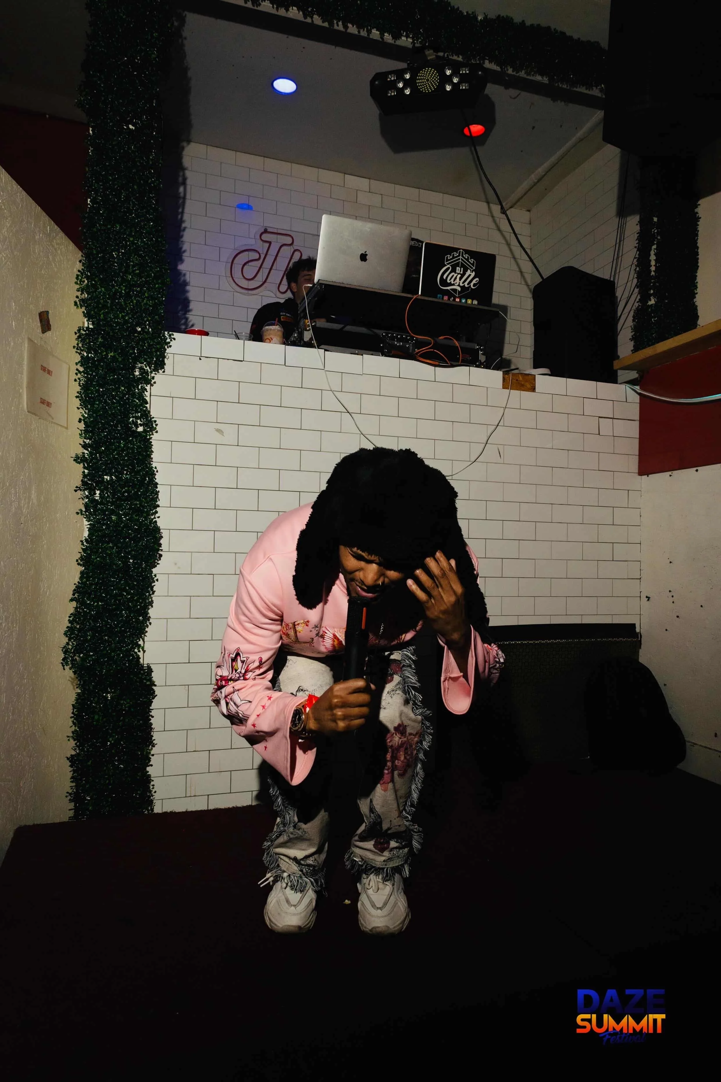 A person in a pink jacket and patterned pants is kneeling on the floor, holding a microphone and covering their face, in a dimly lit setting with a DJ booth and a person behind it in the background. The DJ booth has a keyboard, laptop, and speakers, with colorful lights overhead. There is a sign on the wall with the words "DAZE SUMMIT" in blue and orange fonts.