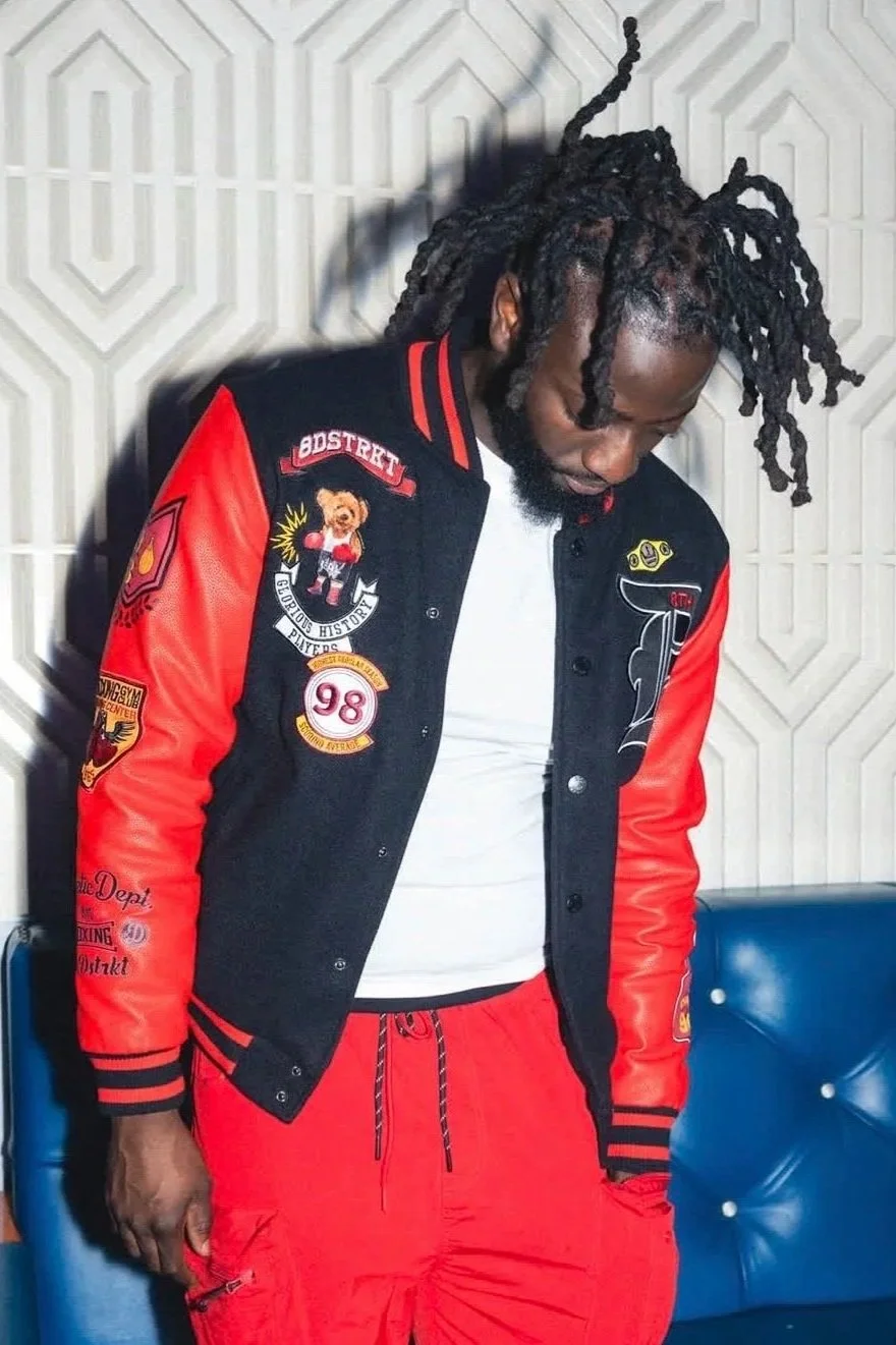 A man with dreadlocks, wearing a black and red varsity jacket with patches, white T-shirt, and red pants, standing against a textured wall.