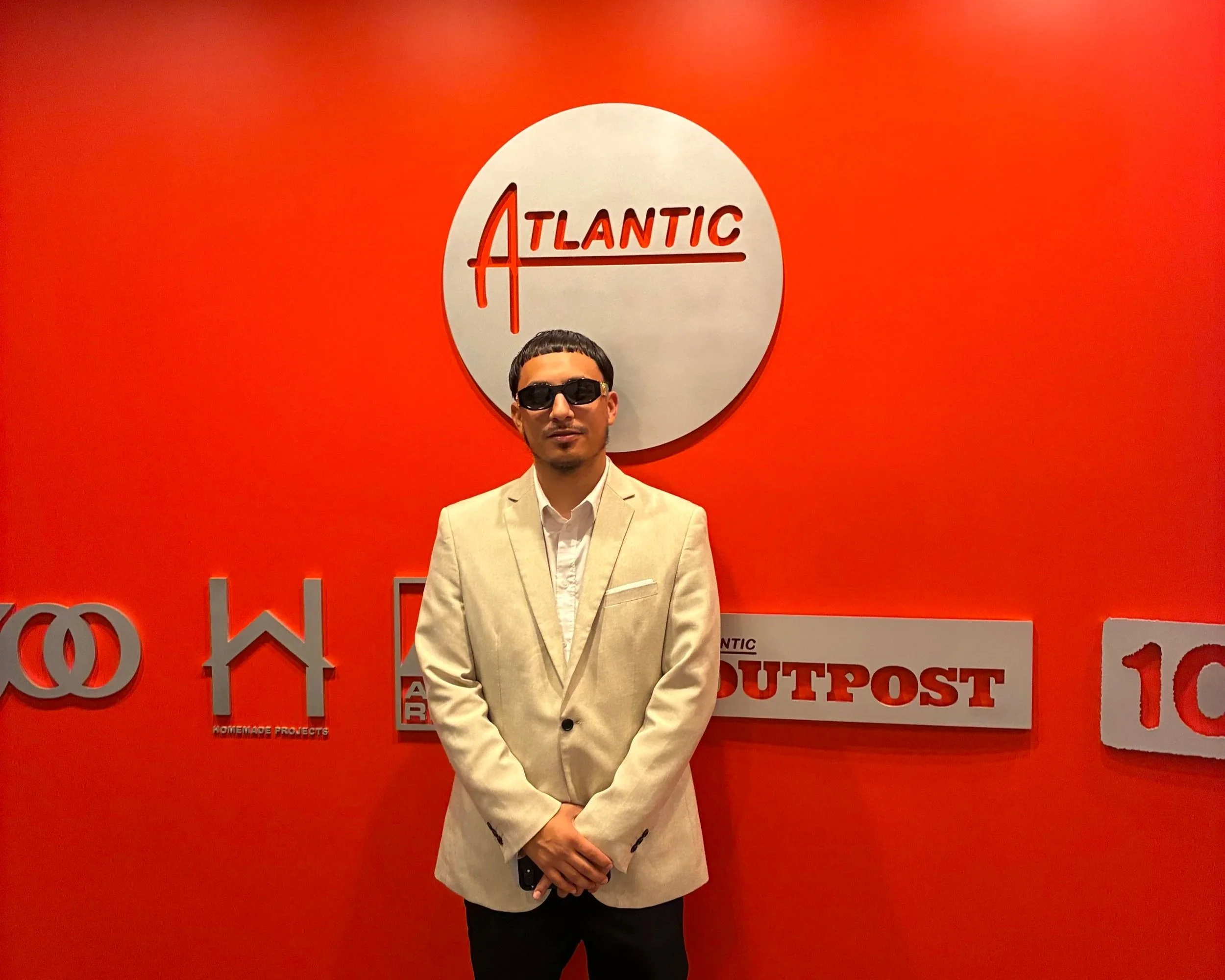 Man wearing sunglasses and a light-colored blazer standing in front of a red wall with the Atlantic logo and text that reads 'HOME MADE PROJECTS' and 'OUTPOST'.
