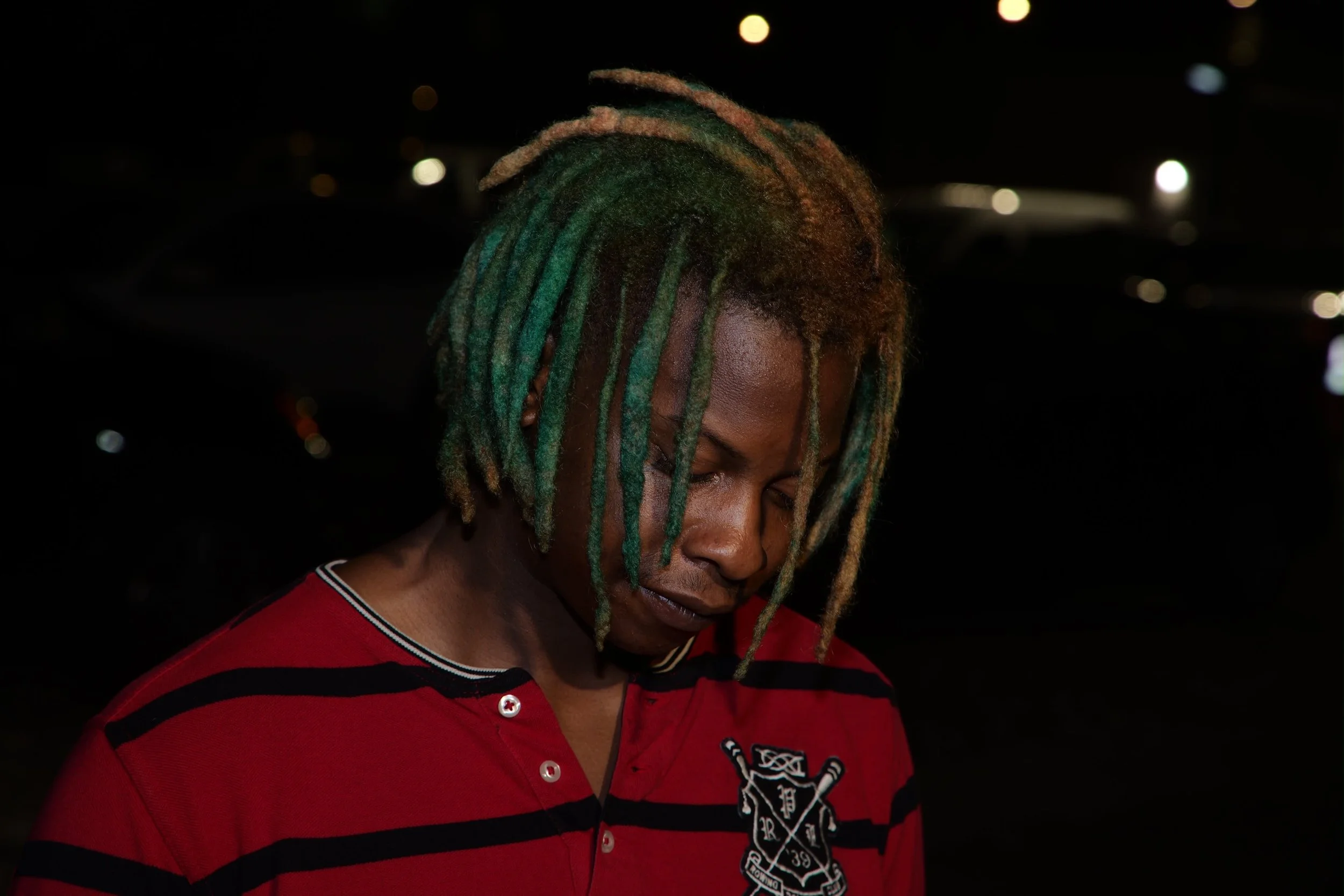 A person with dreadlocks dyed in green and blonde, wearing a red and black striped shirt with an emblem on the chest, shown in a dark environment with blurred lights in the background.