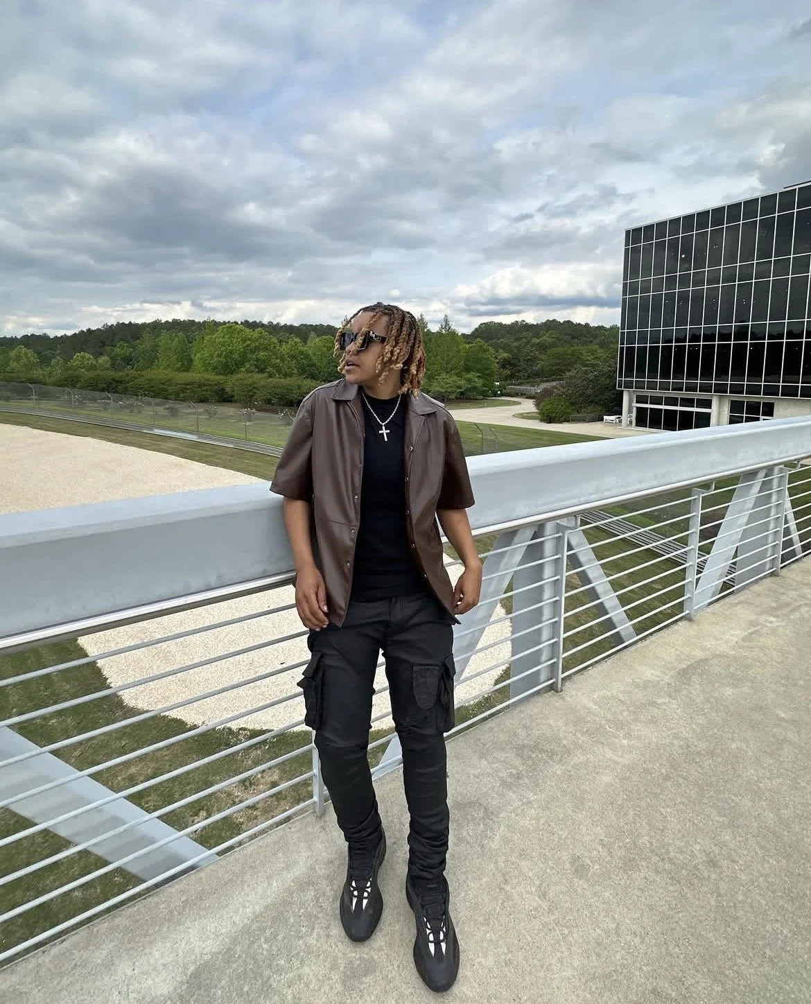 A person with dreadlocks wearing sunglasses, a black shirt, a brown short-sleeve button-up shirt, black cargo pants, and sneakers, standing on a modern pedestrian bridge with metal railings, overlooking a grassy landscape with trees and a modern glass building in the background under cloudy skies.