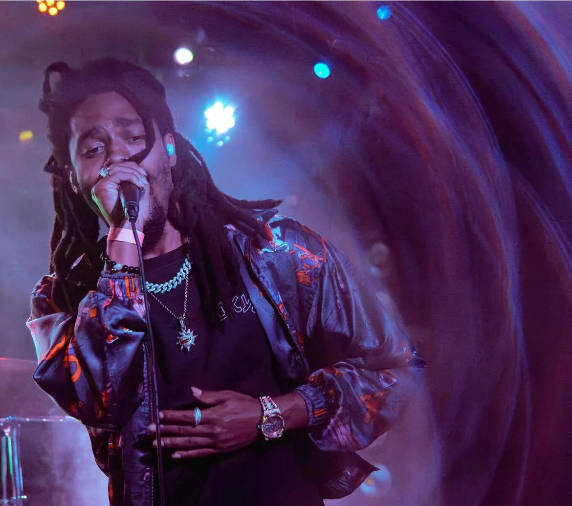 A male performer with dreadlocks singing into a microphone on stage with colorful lighting and a blurred multicolored background.