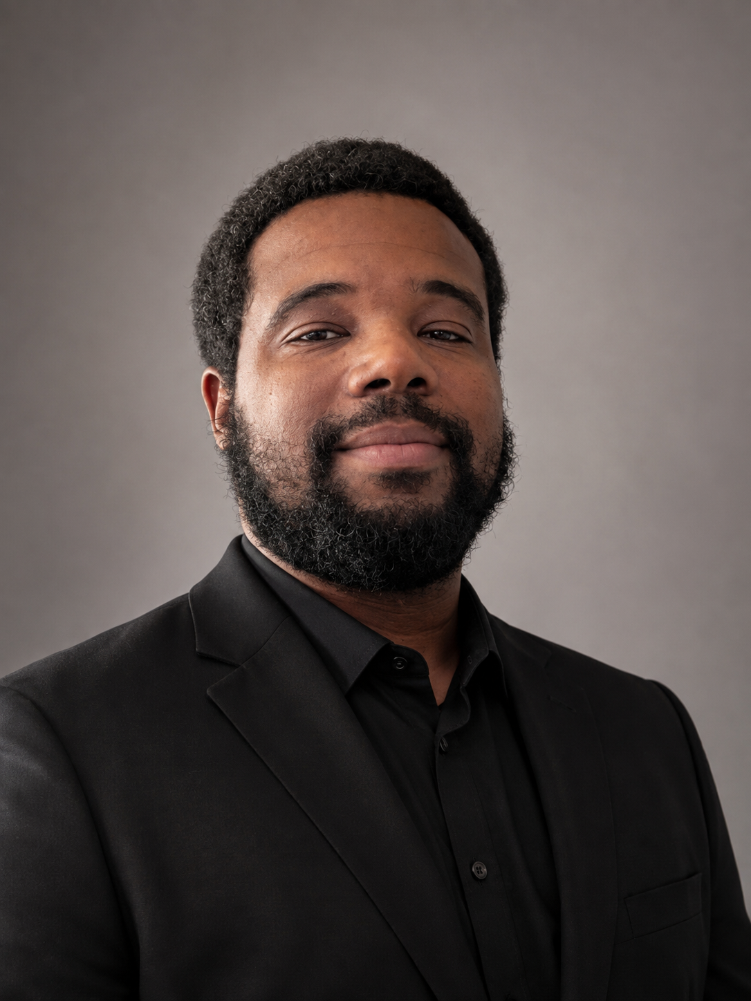 A man with a dark beard and short curly hair wearing a black suit and black shirt against a plain gray background.
