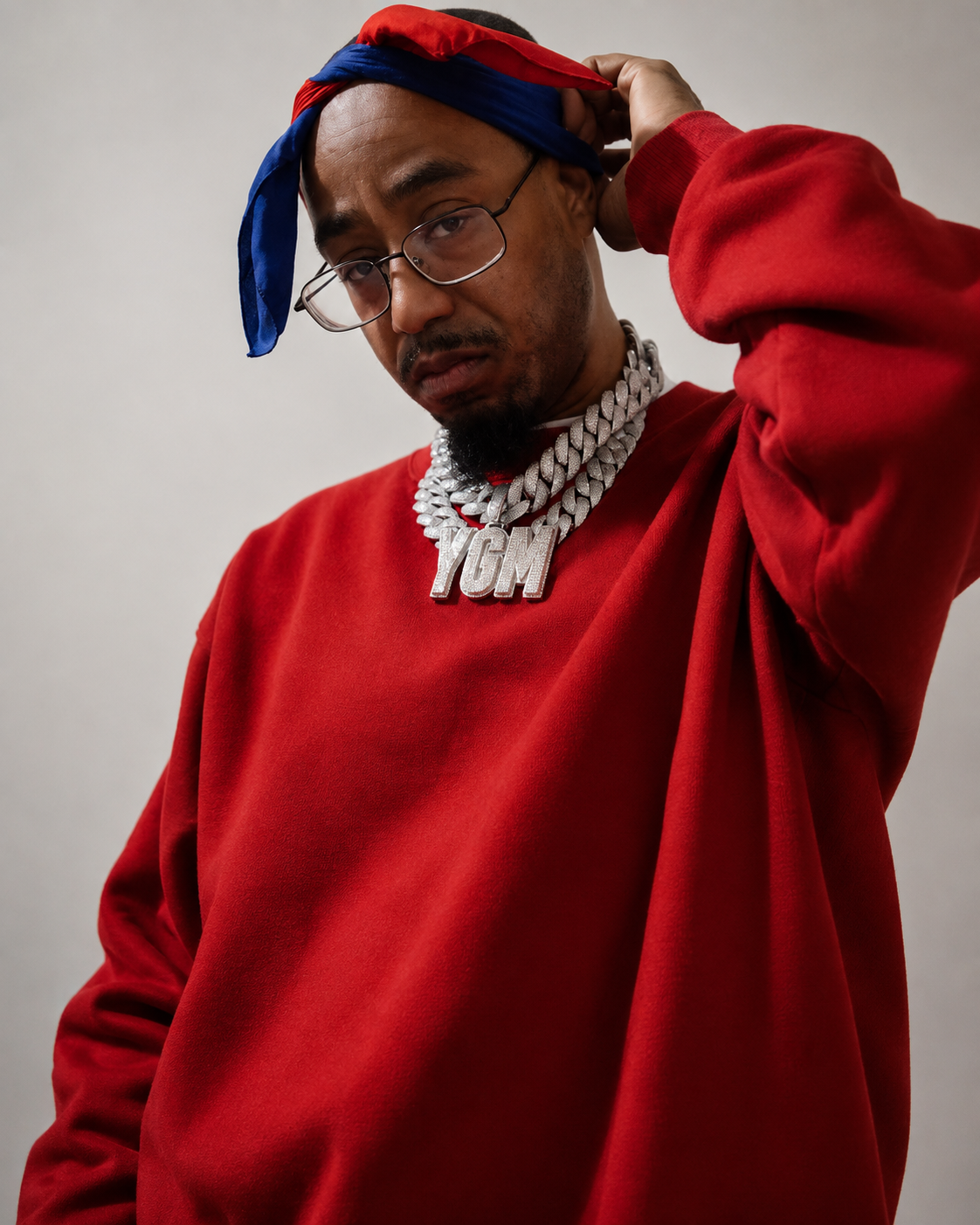 A man wearing a red sweatshirt and glasses, with a red and blue bandana on his head, holding it with his right hand. He has a large silver chain with "YGM" on it around his neck.