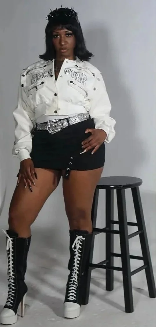 Woman standing next to a black stool, wearing a white jacket, black mini skirt, and high-heeled boots with laces.