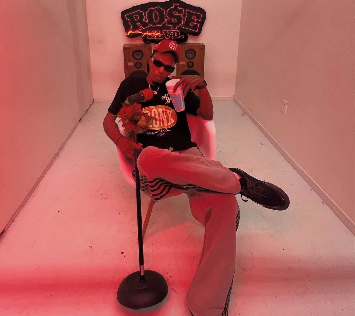 A man wearing sunglasses, a red cap, and casual clothes sitting in a white chair in a room with pink lighting. He is holding a bouquet of flowers in one hand and a drink in the other. Behind him, there is a wall with a neon sign that says 'ROSE' and some speakers.