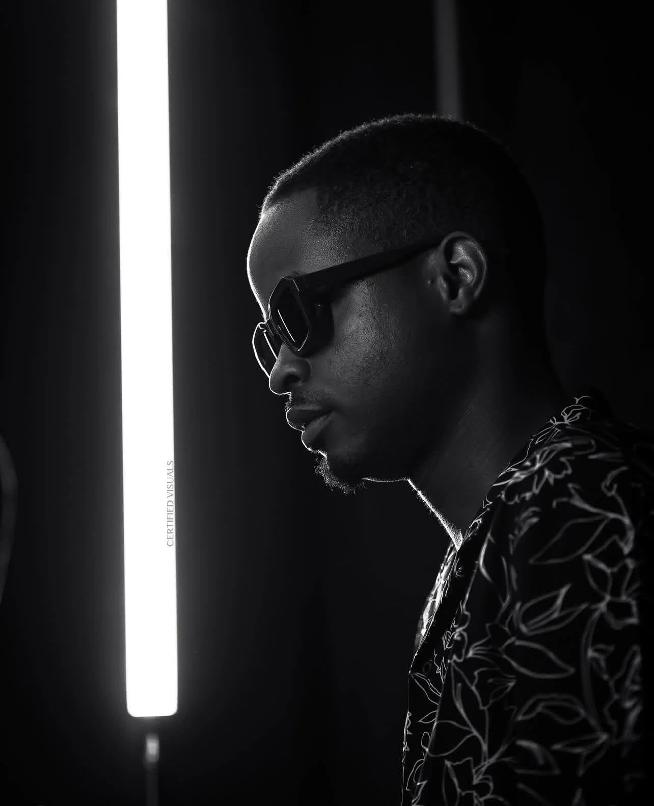 Black and white photo of a man wearing glasses and patterned shirt, standing beside a vertical bright light in a dark room.