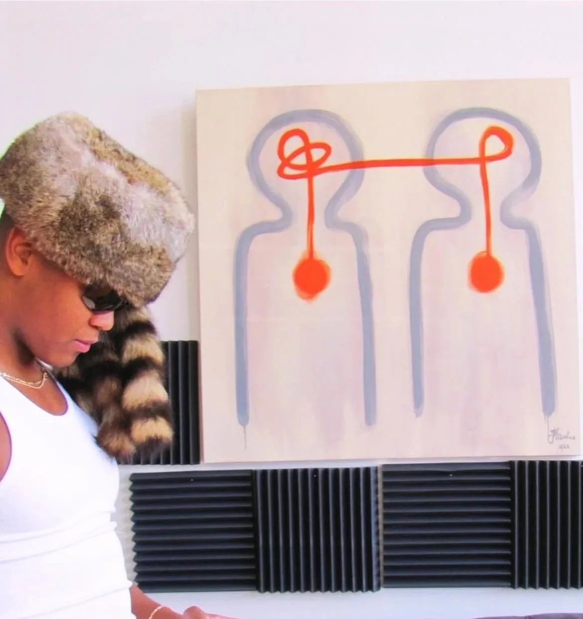 Person wearing a large fur hat, sunglasses, and a white tank top, looking down. There is abstract art on the wall featuring two stylized human outlines with orange dots and lines connecting them, and black acoustic foam panels below.