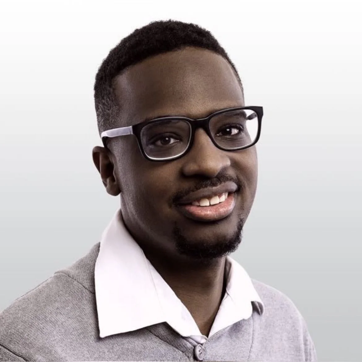 Portrait of a young man with glasses, smiling, wearing a white collared shirt and gray sweater, against a neutral background.