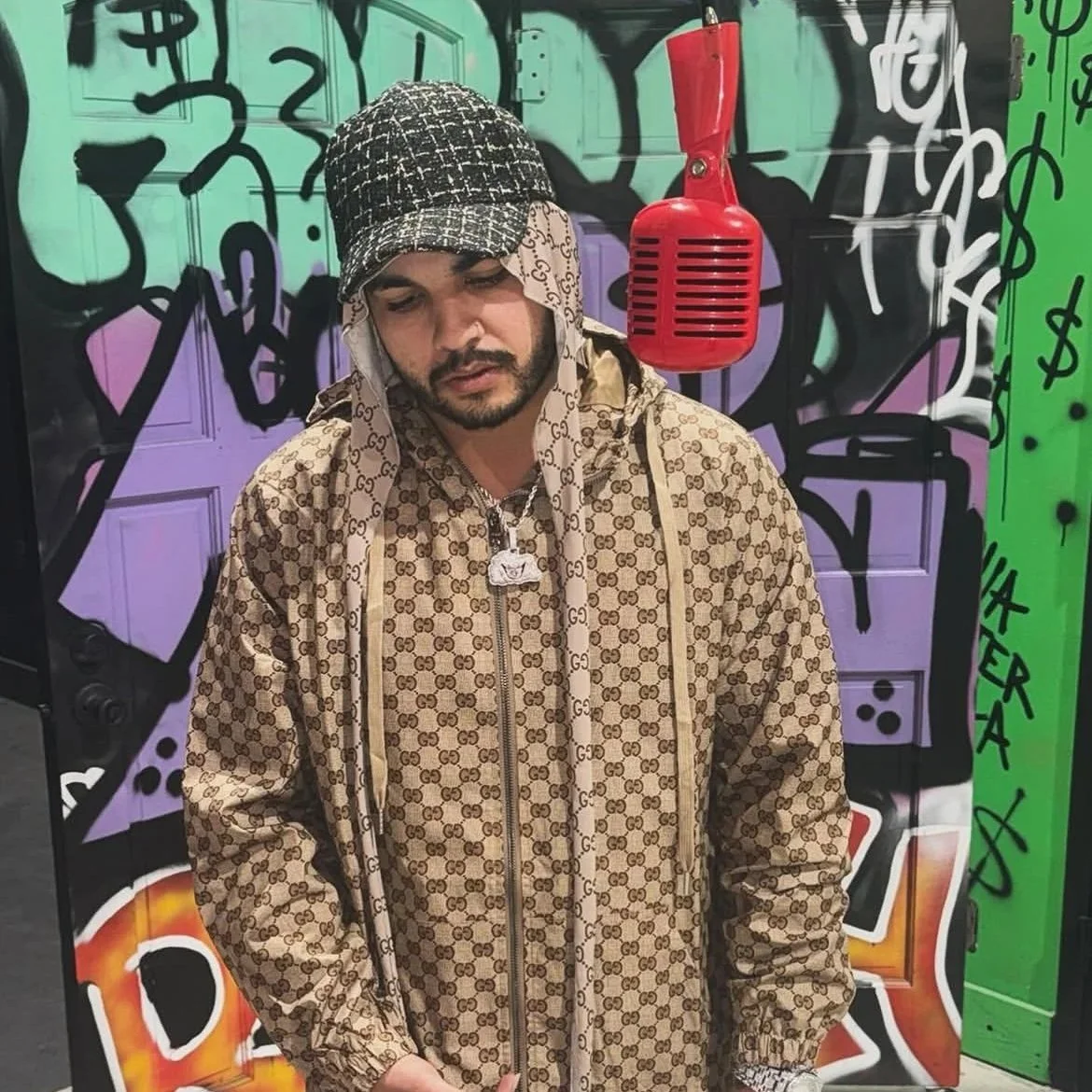 Man wearing a Gucci patterned hoodie and a Burberry patterned bandana standing in front of graffiti art and a red microphone.