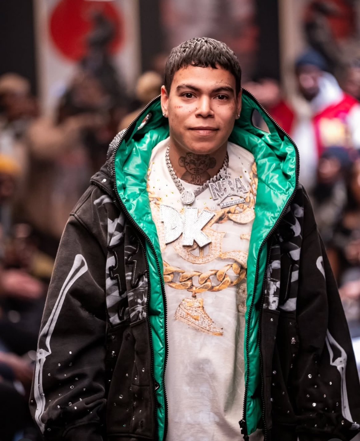 Young man with short dark hair, wearing a white T-shirt with the Nike logo adorned with jewelry, including chains and necklaces, a black and gray jacket with a green lining, and earrings, standing in a crowded area.