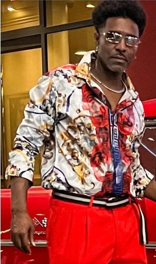 Man with an afro hairstyle wearing sunglasses, a colorful patterned shirt, red pants, and a striped belt, standing in front of a classic car with a red exterior inside a building with yellow and red walls.