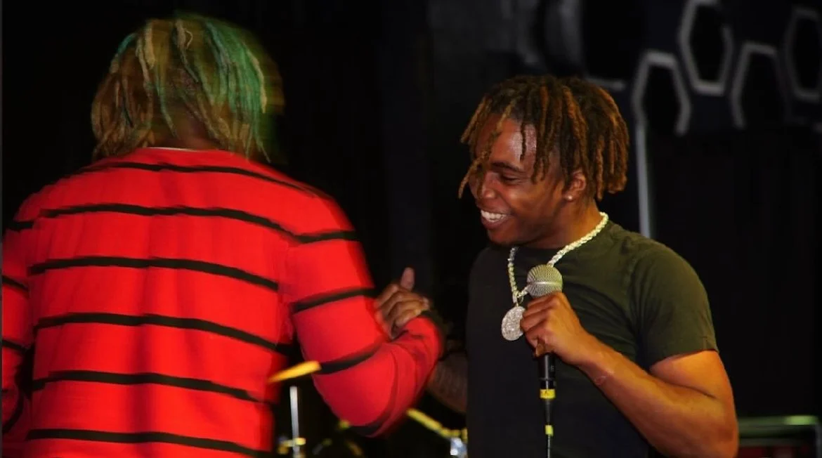 A man with dreadlocks smiling and holding a microphone on stage, facing another person with dreadlocks and wearing a red and black striped shirt.