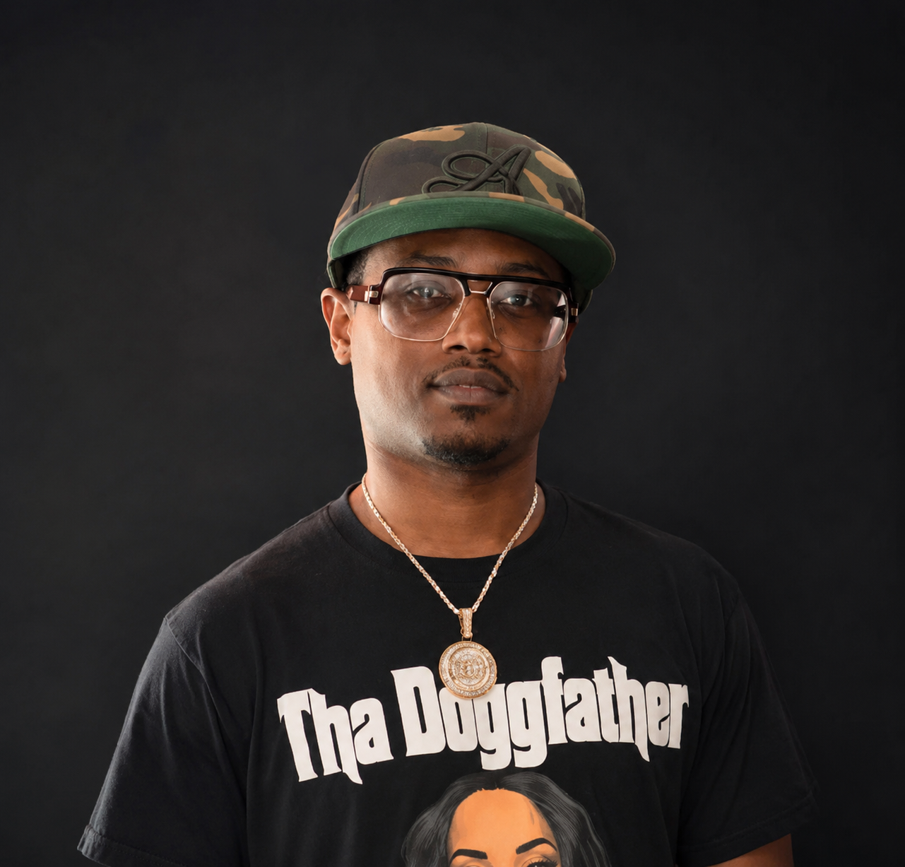 A man wearing glasses, a camo cap, a black Thugfather T-shirt, and a gold chain with a medallion, posing against a black background.