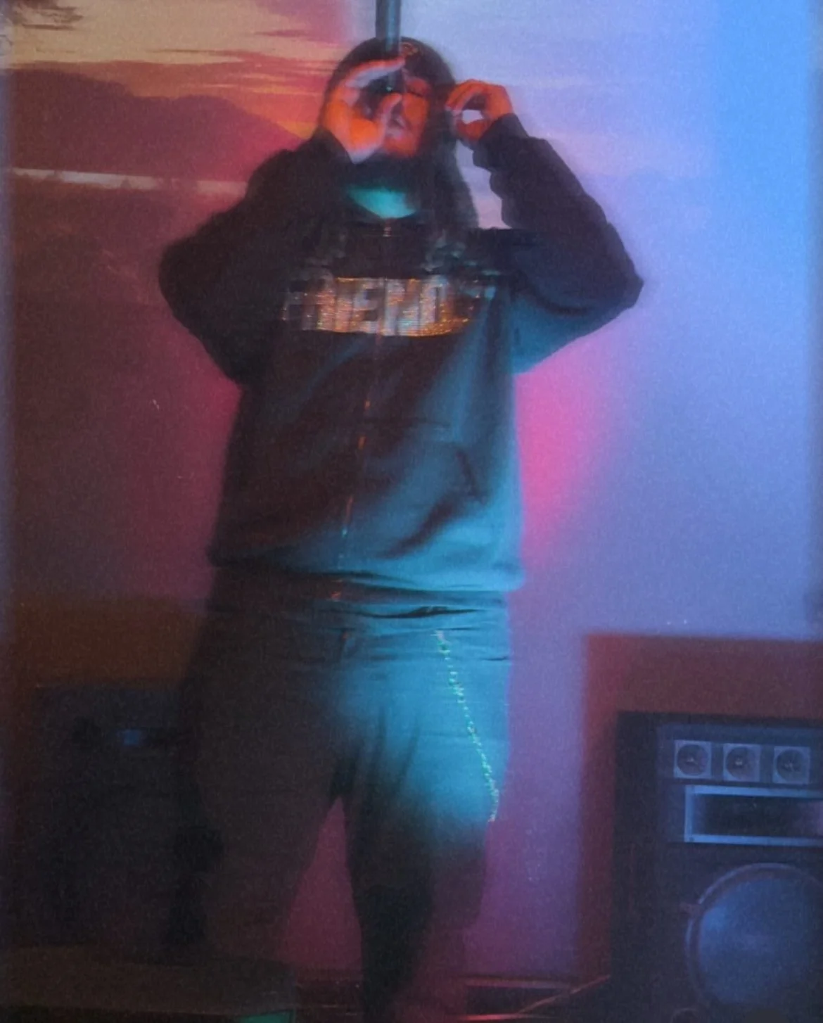 A person taking a selfie in a room with colorful lighting, wearing a hoodie and pants, with a speaker visible in the background.