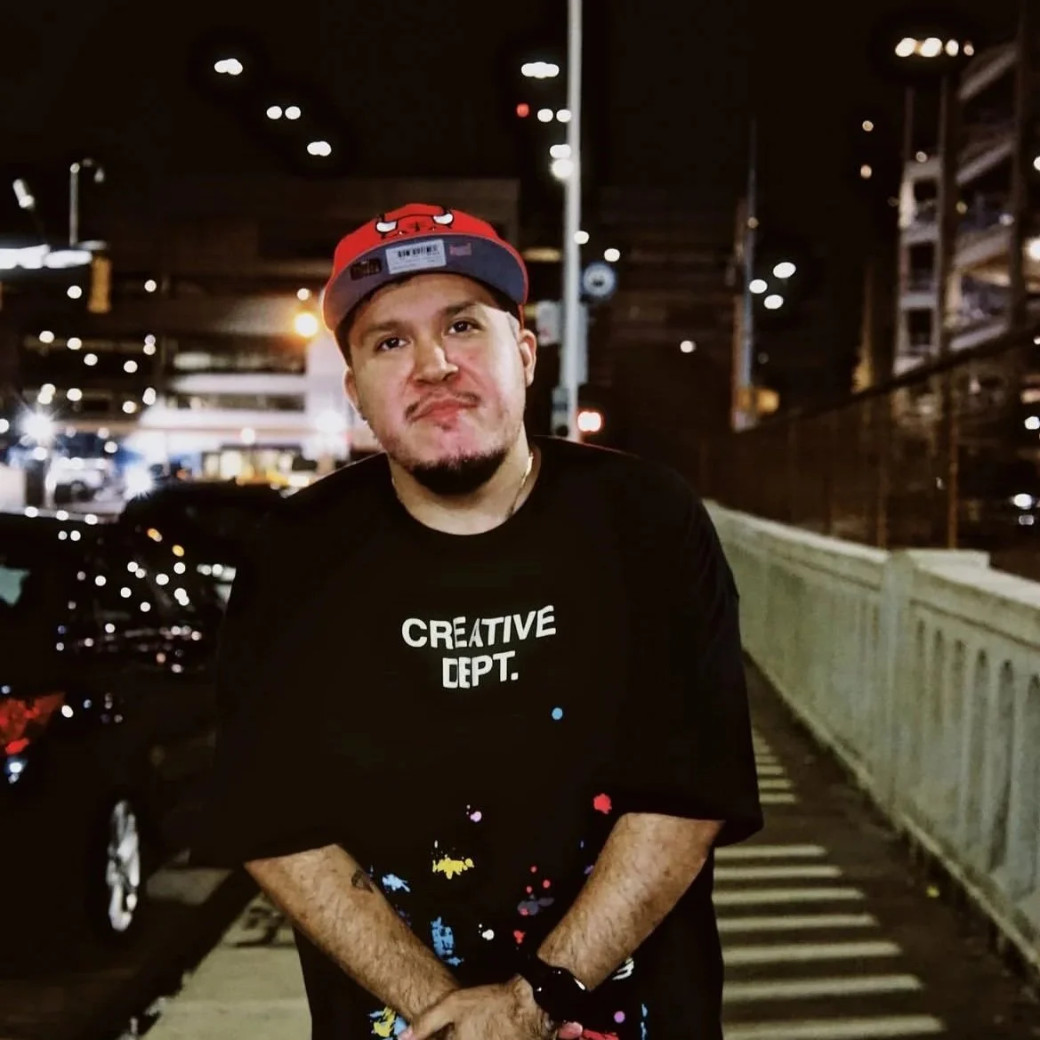 A young man standing on a city sidewalk at night, wearing a red and gray cap with a cartoon character, a black t-shirt with 'CREATIVE DEPT.' printed on it, and a watch. Night city lights and parked cars are visible in the background.