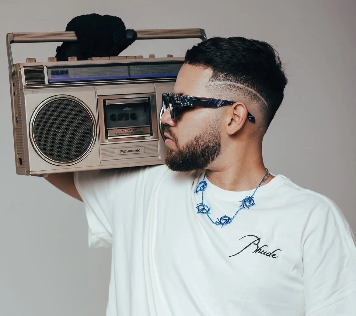Man with beard and styled haircut wearing sunglasses, white t-shirt, and blue necklace, holding a vintage Panasonic boombox on his shoulder, against a plain gray background.