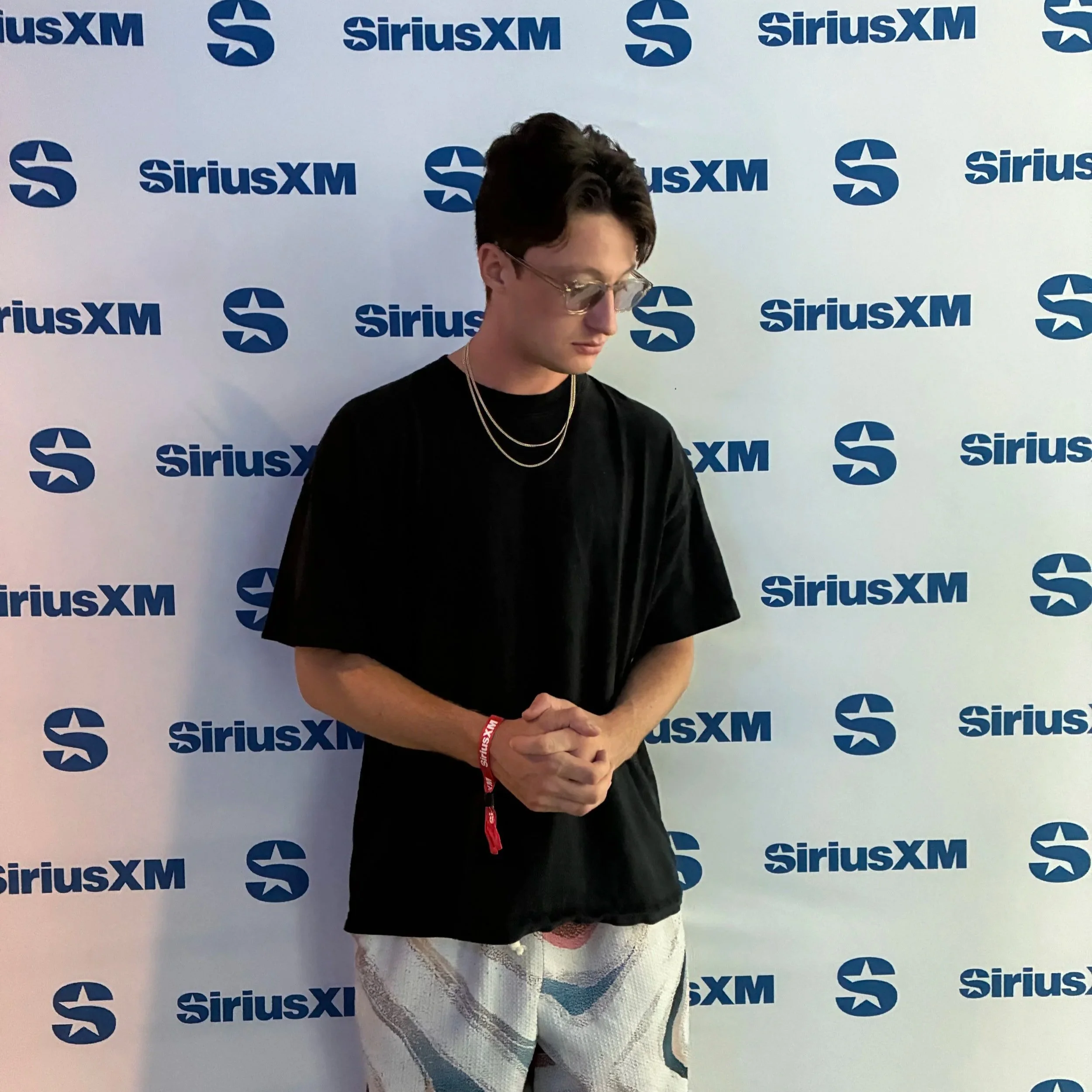 A person with short dark hair, wearing glasses, a black t-shirt, and layered necklaces, standing in front of a white background with blue SiriusXM logos. The person is looking down with hands clasped in front.