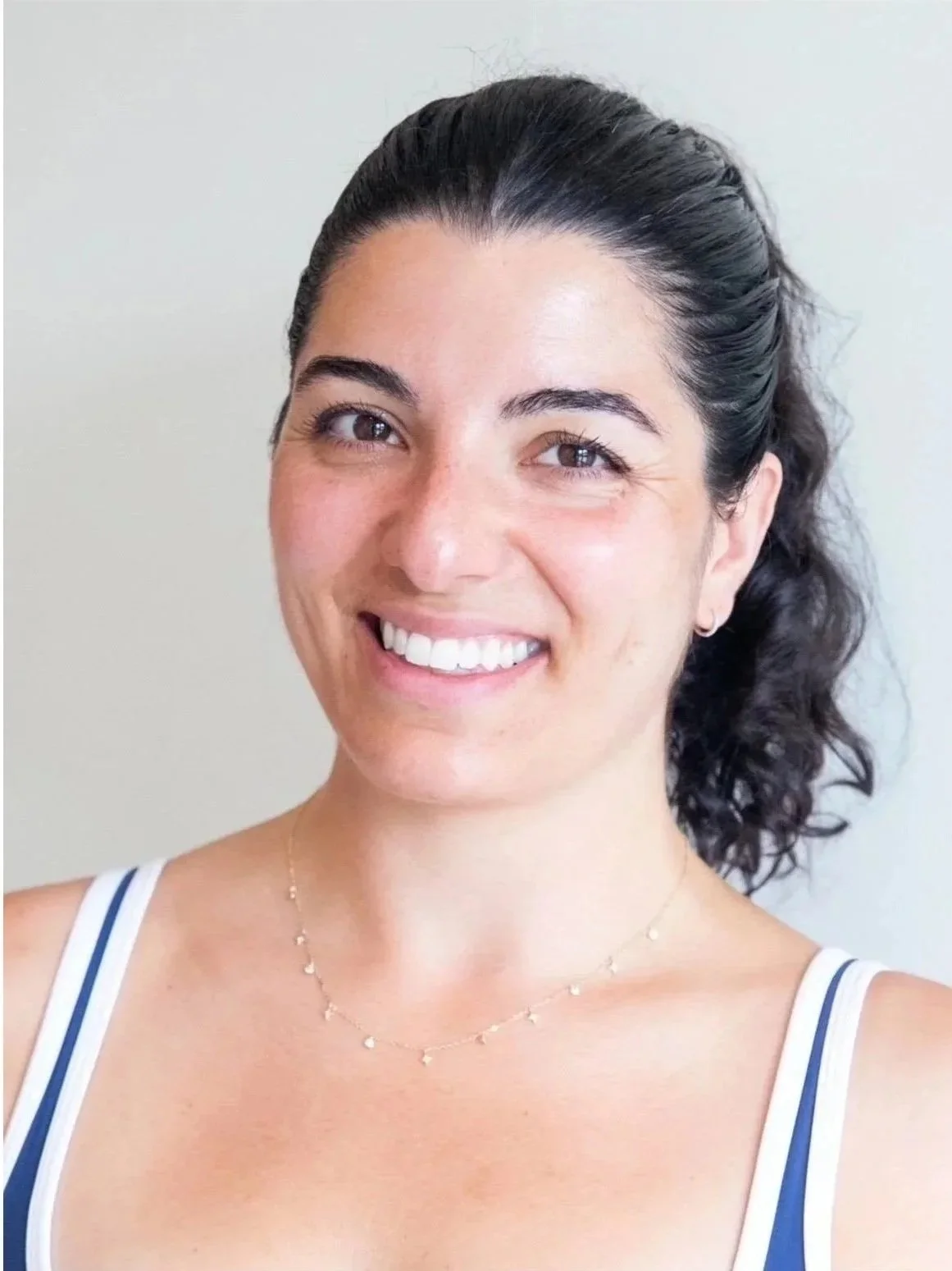 A woman with dark, curly hair pulled back, smiling, wearing a white tank top with blue straps, a delicate gold necklace with small charms, and small earrings, standing against a plain light-colored background.
