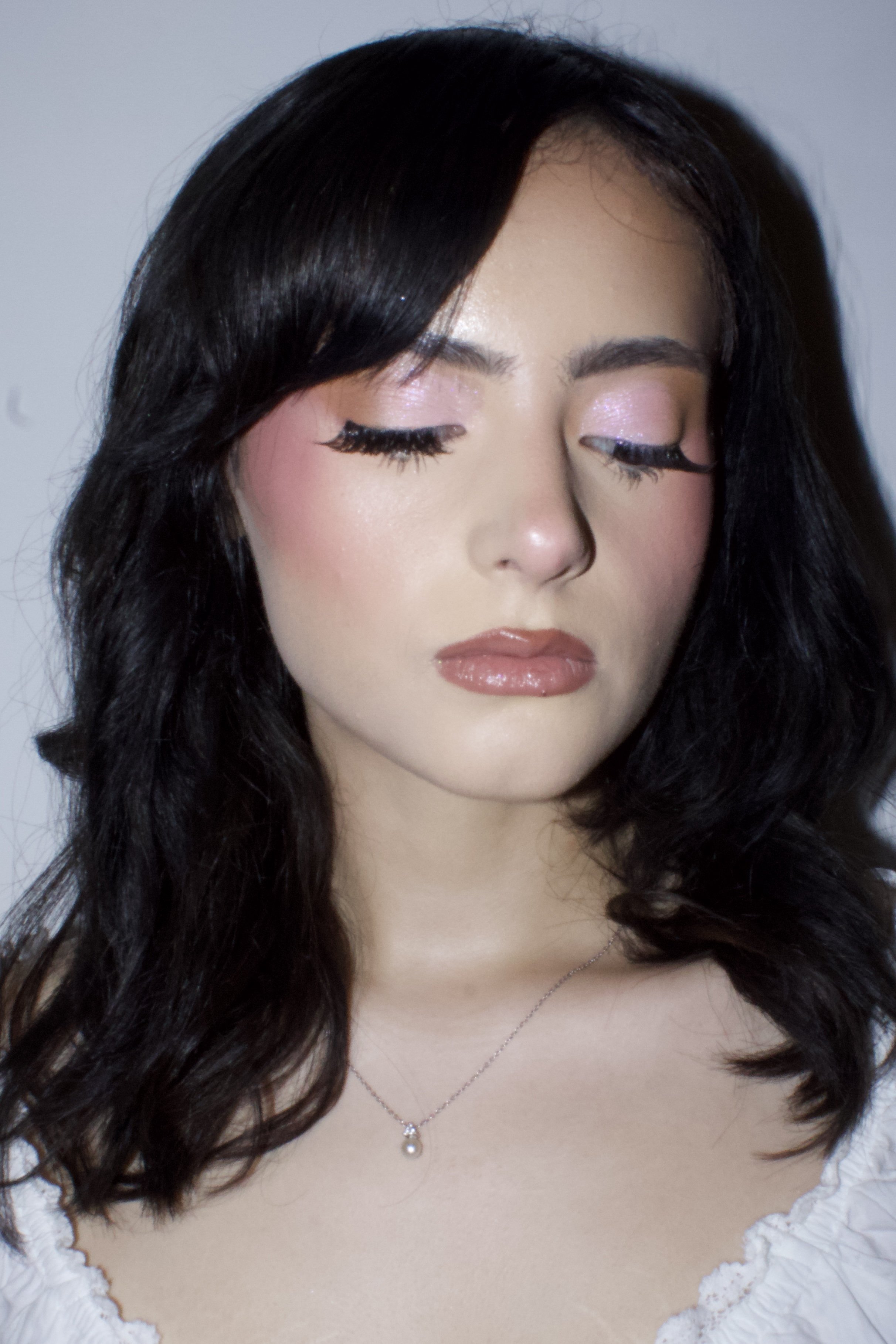 Close-up of a woman with long black hair, closed eyes, pink eyeshadow, and shiny lips, wearing a white lace-top dress and a pearl necklace with a pendant.