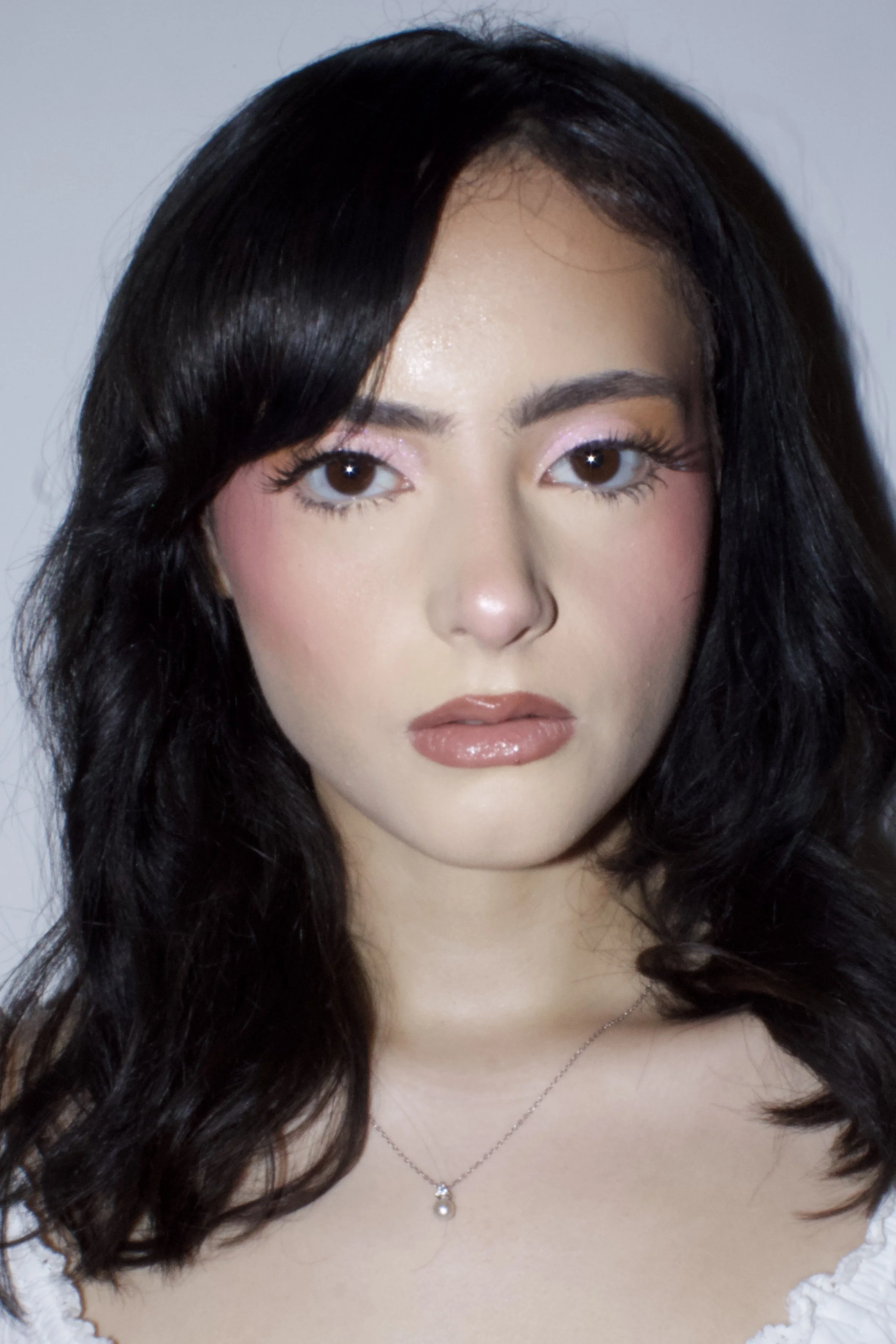 Close-up of a woman with long black hair, wearing pink eyeshadow, blush, lipstick, and a pearl necklace with a small pendant, against a light background.