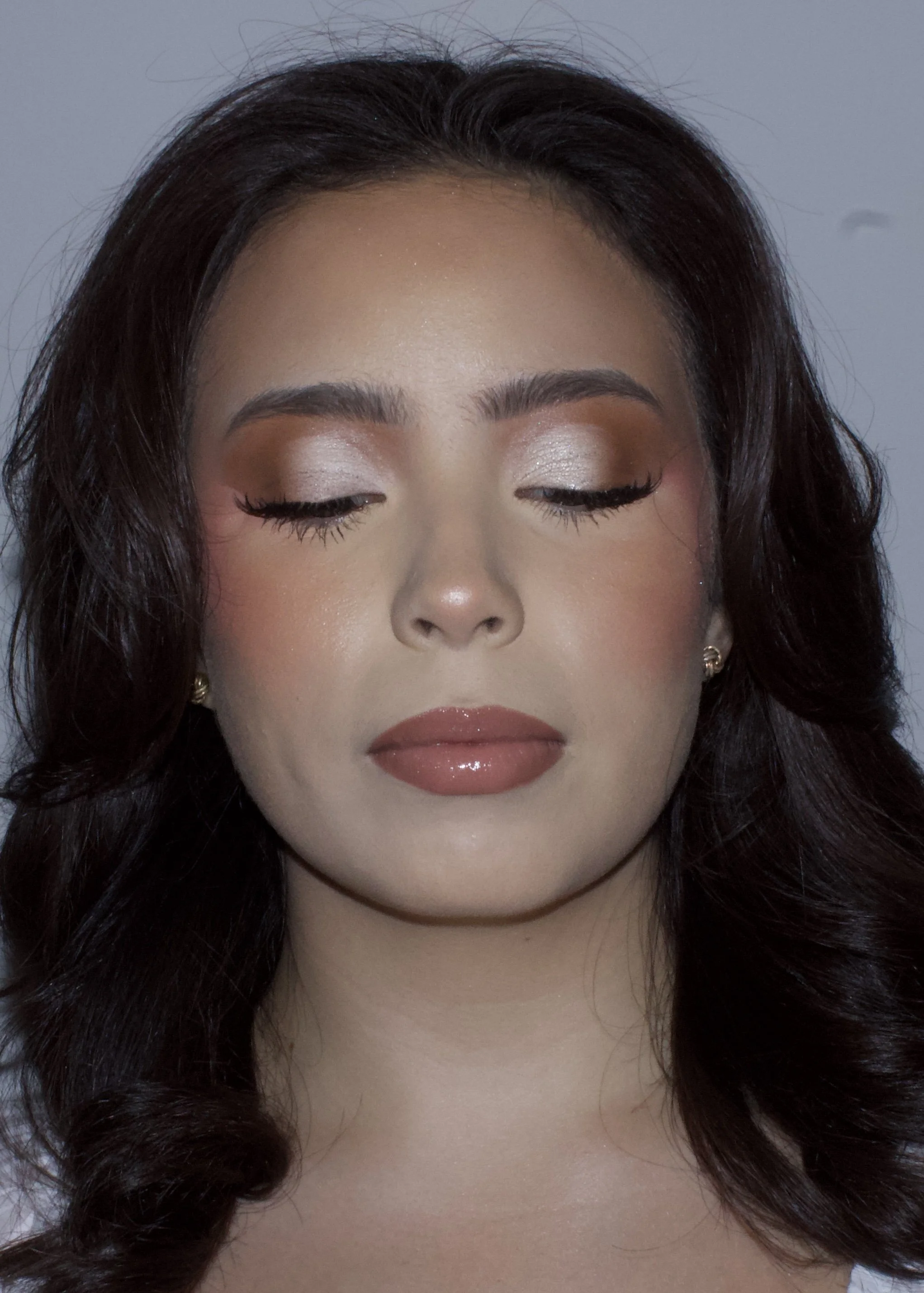 Close-up of a woman with dark wavy hair, makeup including shimmery eyeshadow, eyeliner, mascara, blush, and glossy lips, with a neutral background.