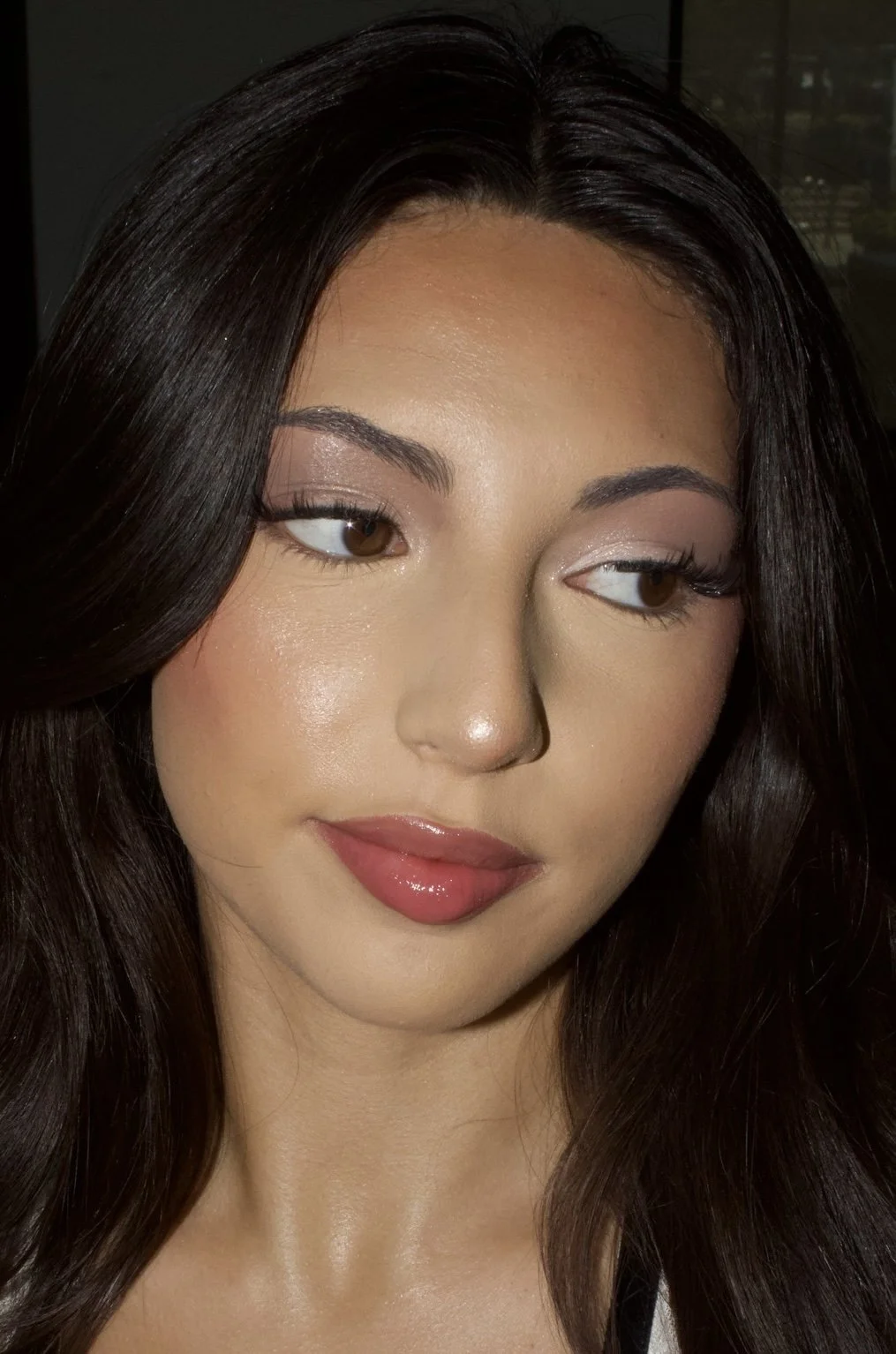A close-up portrait of a woman with long dark hair, brown eyes, makeup, and pink lipstick, looking slightly to the side with a soft smile.