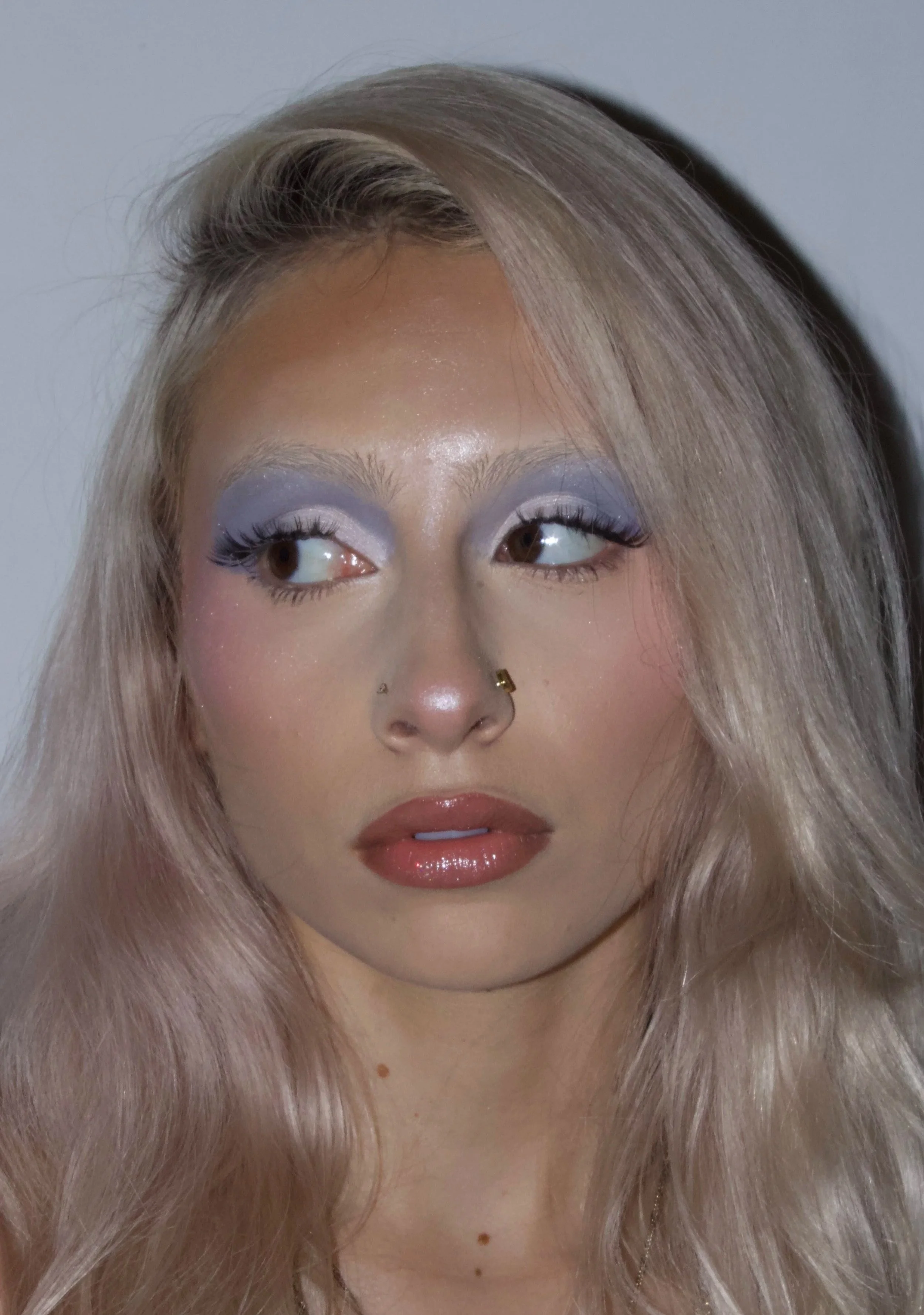 Portrait of a woman with long, blonde hair and makeup including purple eyeshadow, mascara, and glossy lips, against a plain background.